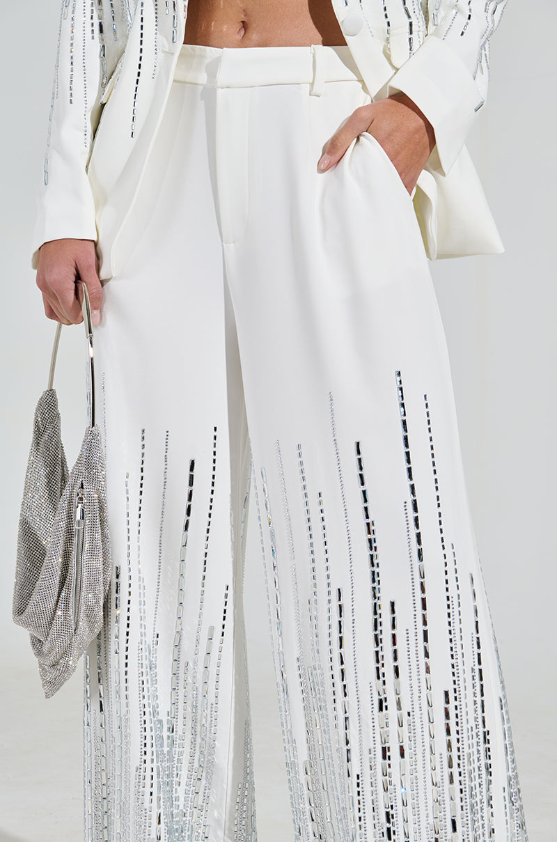 Someone is wearing a stylish white suit with the MILA EMBELLISHED FLARE PANT, high-rise wide-leg pants adorned with sequins in vertical patterns. They hold a silver, glittery clutch, adding a touch of elegance and glamour.