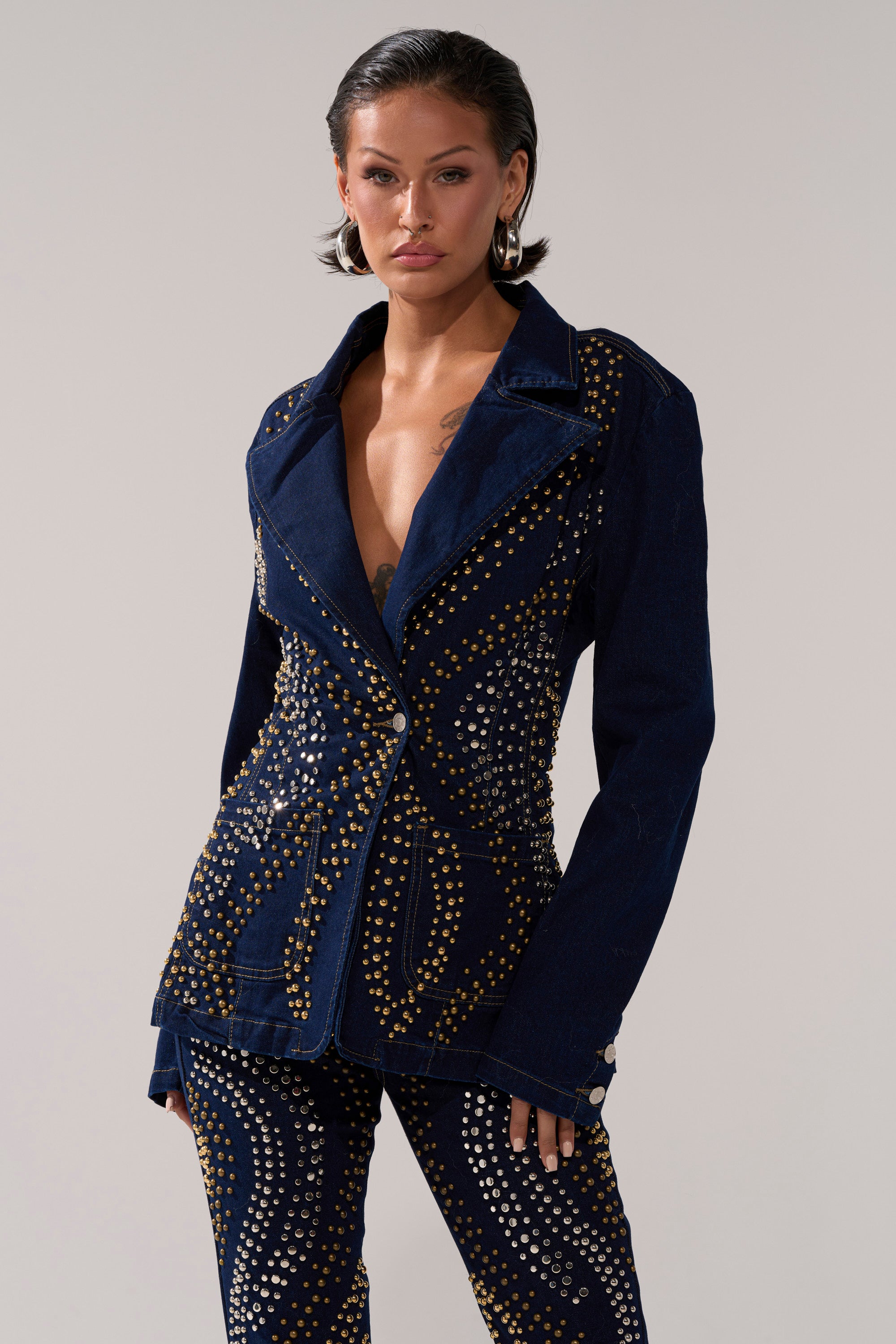 A woman with short dark hair confidently models the CHEYENNE STUDDED DENIM BLAZER and matching pants, featuring gold and silver stud details, against a plain light background.