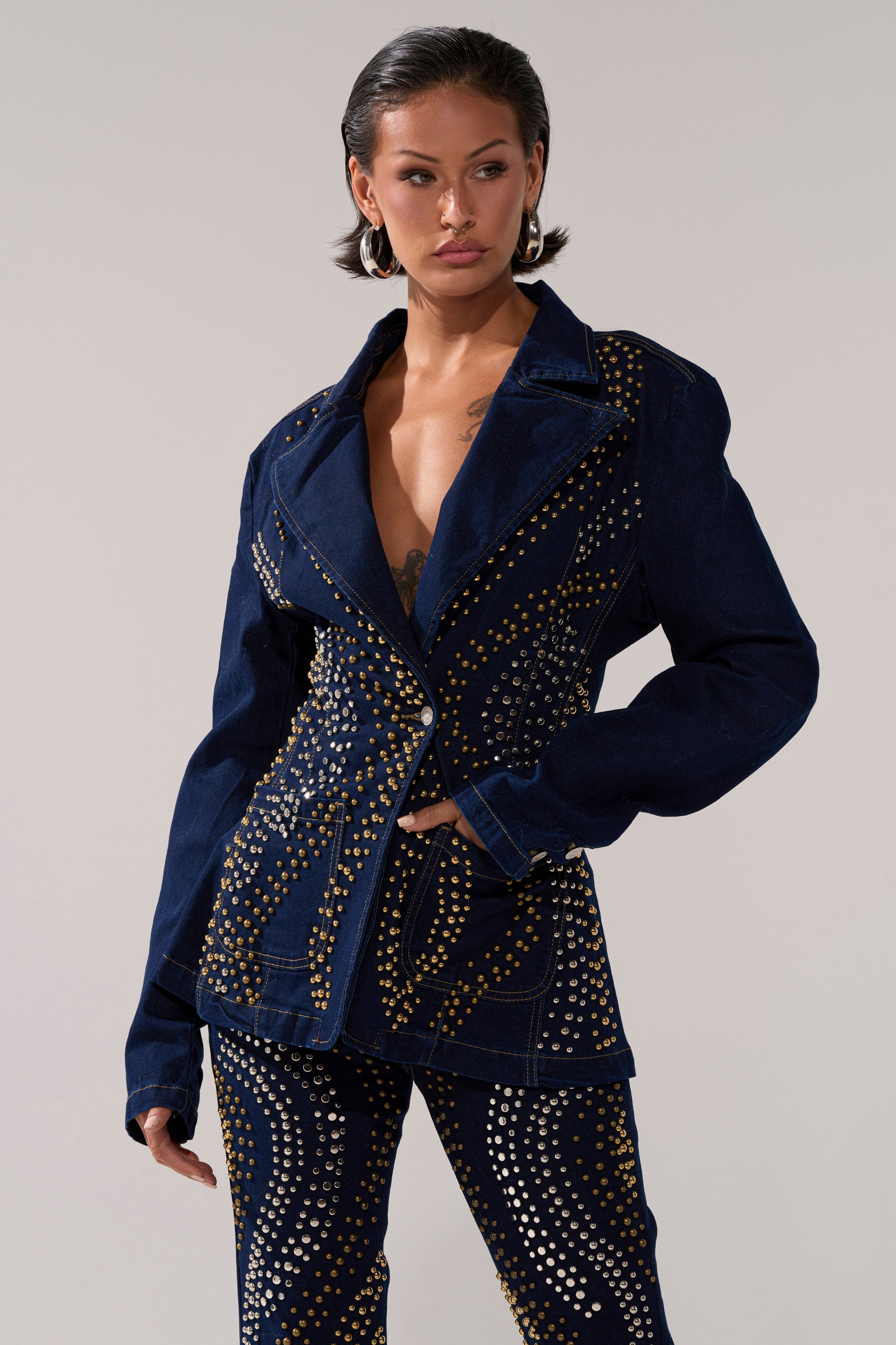 A woman wearing the CHEYENNE STUDDED DENIM BLAZER, a dark-wash piece adorned with swirling gold studs and paired with matching pants, stands against a plain backdrop with her hand on her hip, gazing to the side.