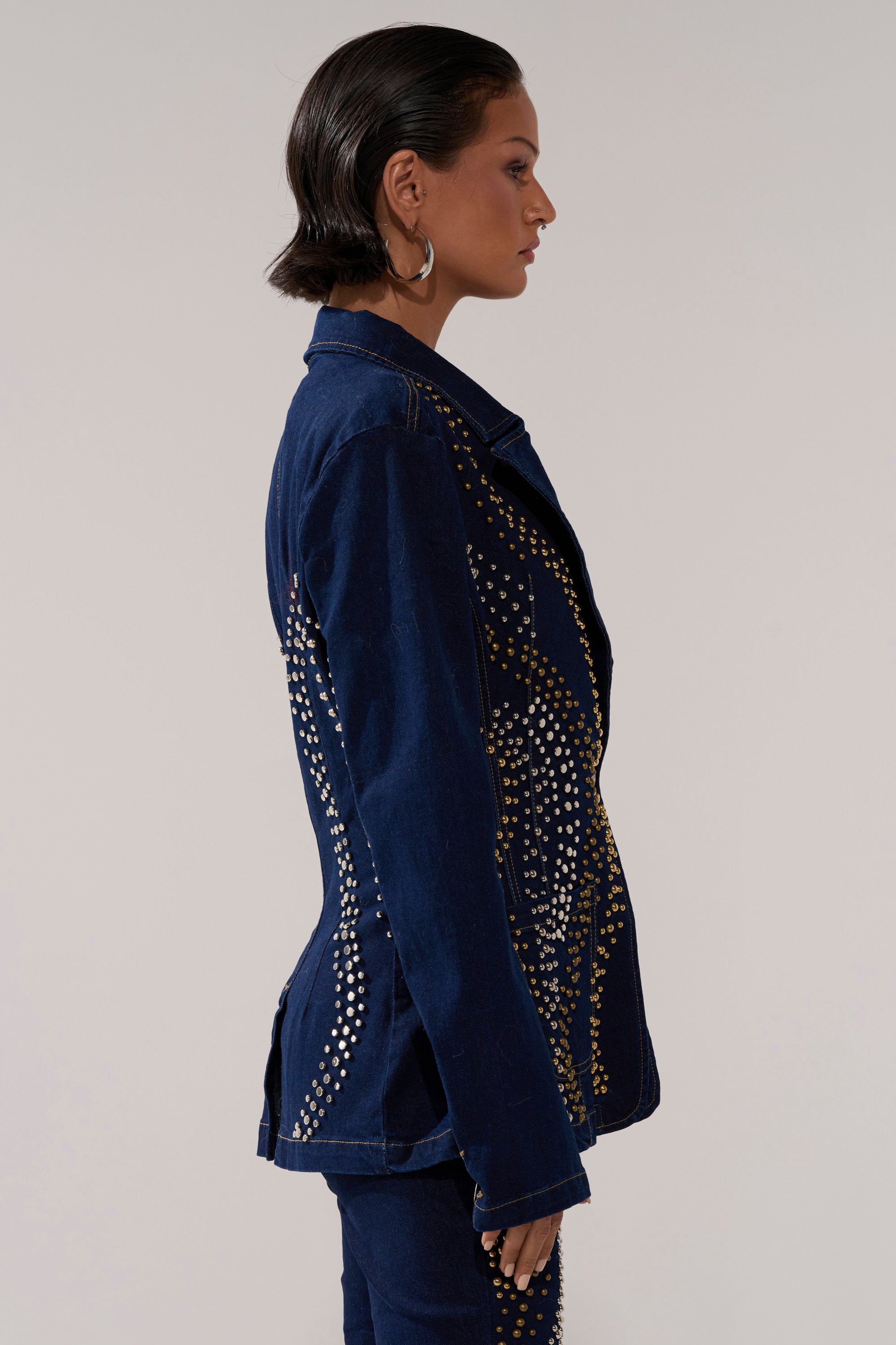 A person with short, slicked-back hair models the CHEYENNE STUDDED DENIM BLAZER, featuring silver and gold studs, viewed in profile against a plain light background.