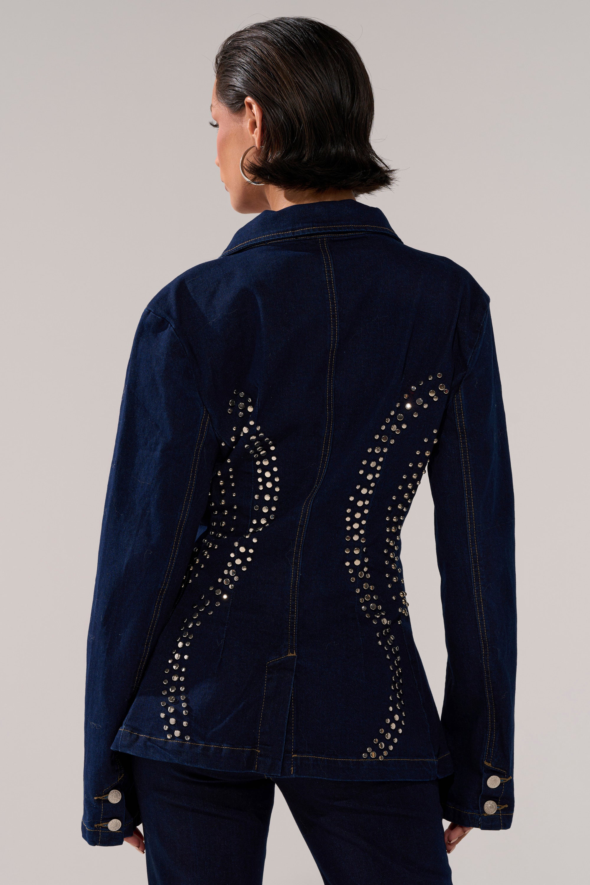 A person with short dark hair and hoop earrings stands facing away from the camera, wearing the CHEYENNE STUDDED DENIM BLAZER featuring silver studs in curved patterns on the back, against a neutral background.