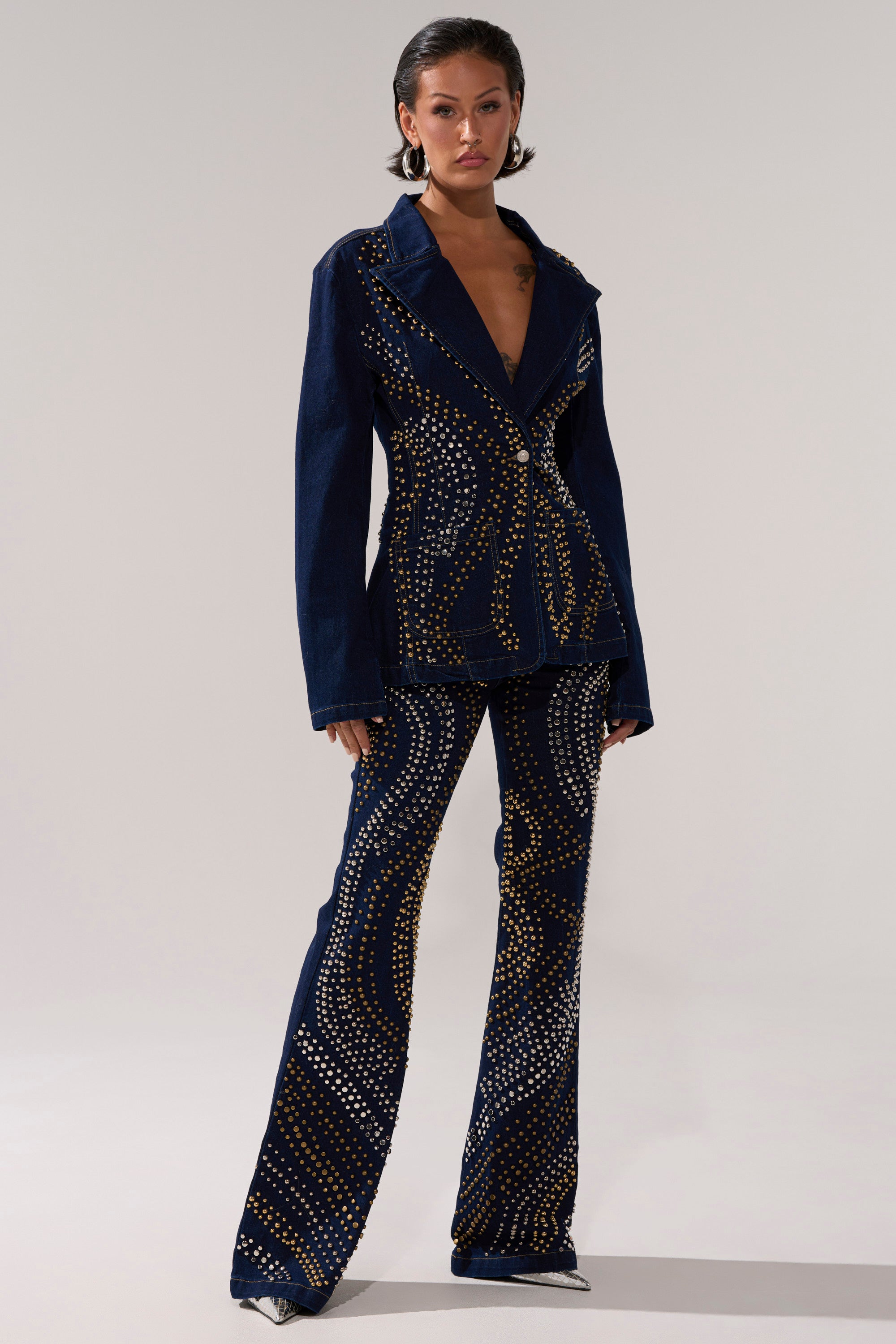 A person stands confidently in the CHEYENNE STUDDED DENIM BLAZER and flared pants, featuring gold and silver dot patterns with geometric, swirling designs. They complete the look with pointed silver shoes and short dark hair.