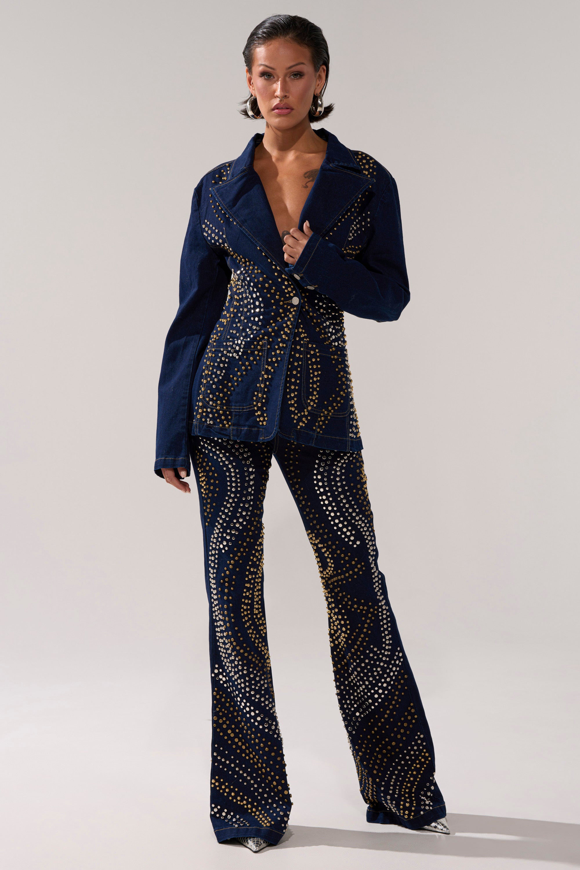 A person stands confidently in the CHEYENNE STUDDED DENIM BLAZER, a navy blue piece with gold studs and silver swirling embellishments, fitted silhouette, and paired with matching flared pants against a plain light background.