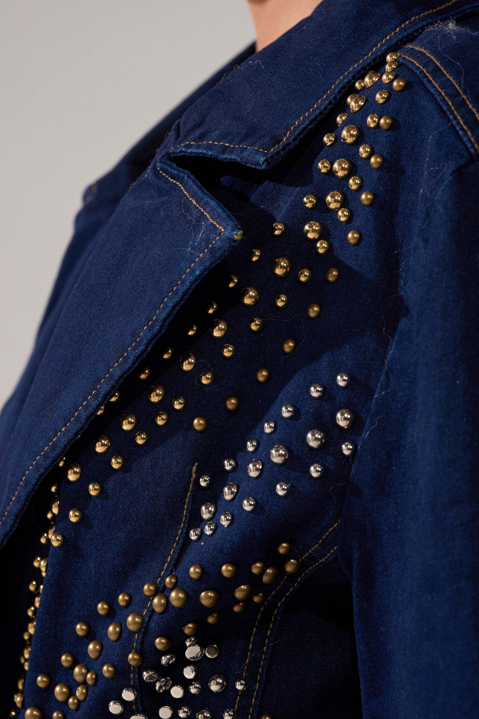 Close-up of the CHEYENNE STUDDED DENIM BLAZER in dark wash, featuring gold and silver studs in clustered patterns, detailed stitching, and a glimpse of the collar.