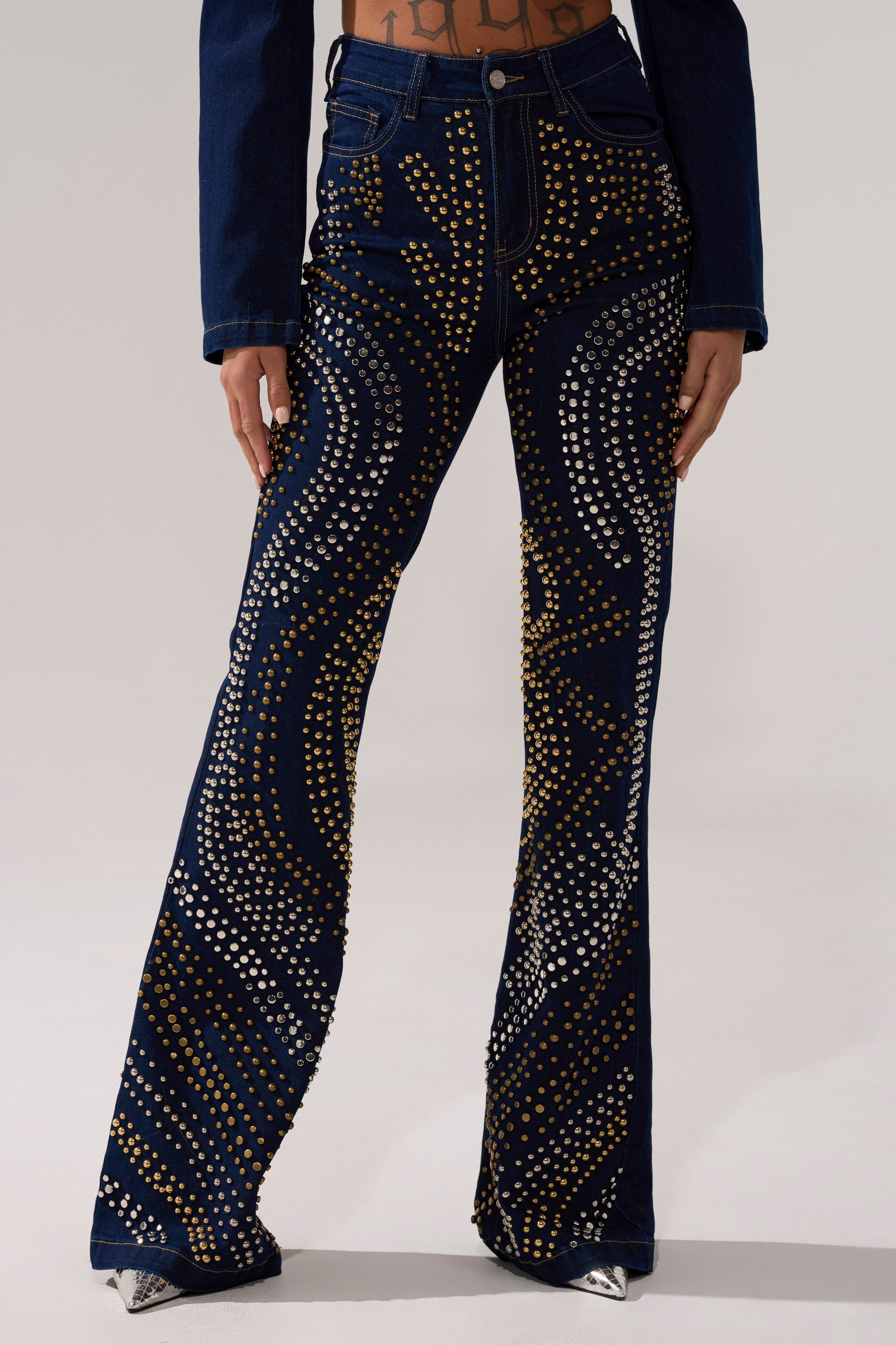 Wearing CHEYENNE STUDDED FLARE DENIM, featuring high-rise denim pants with gold and silver circular studs. Only the lower torso and legs are visible, showcasing the unique studded flare style.