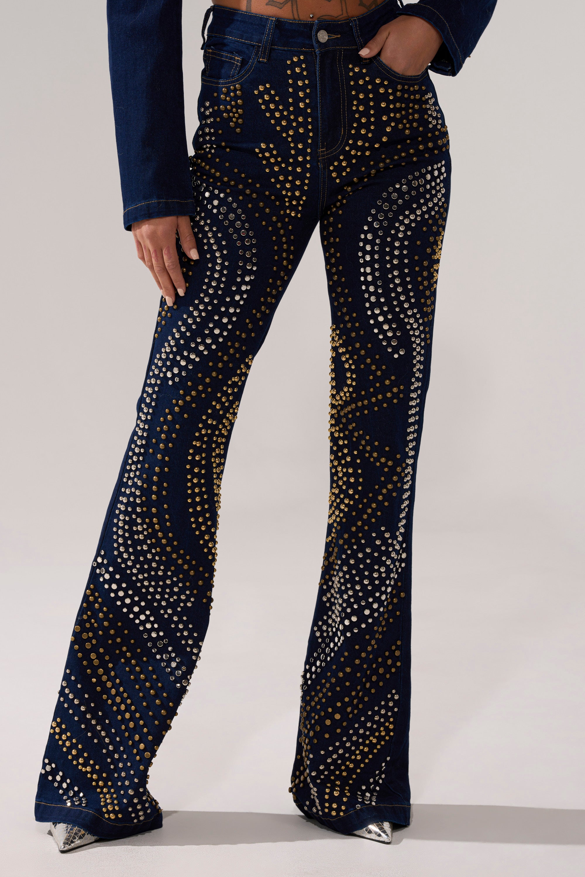 Wearing the CHEYENNE STUDDED FLARE DENIM—gold and silver studded flared jeans with swirling patterns—paired with a matching denim jacket and metallic pointed-toe shoes.