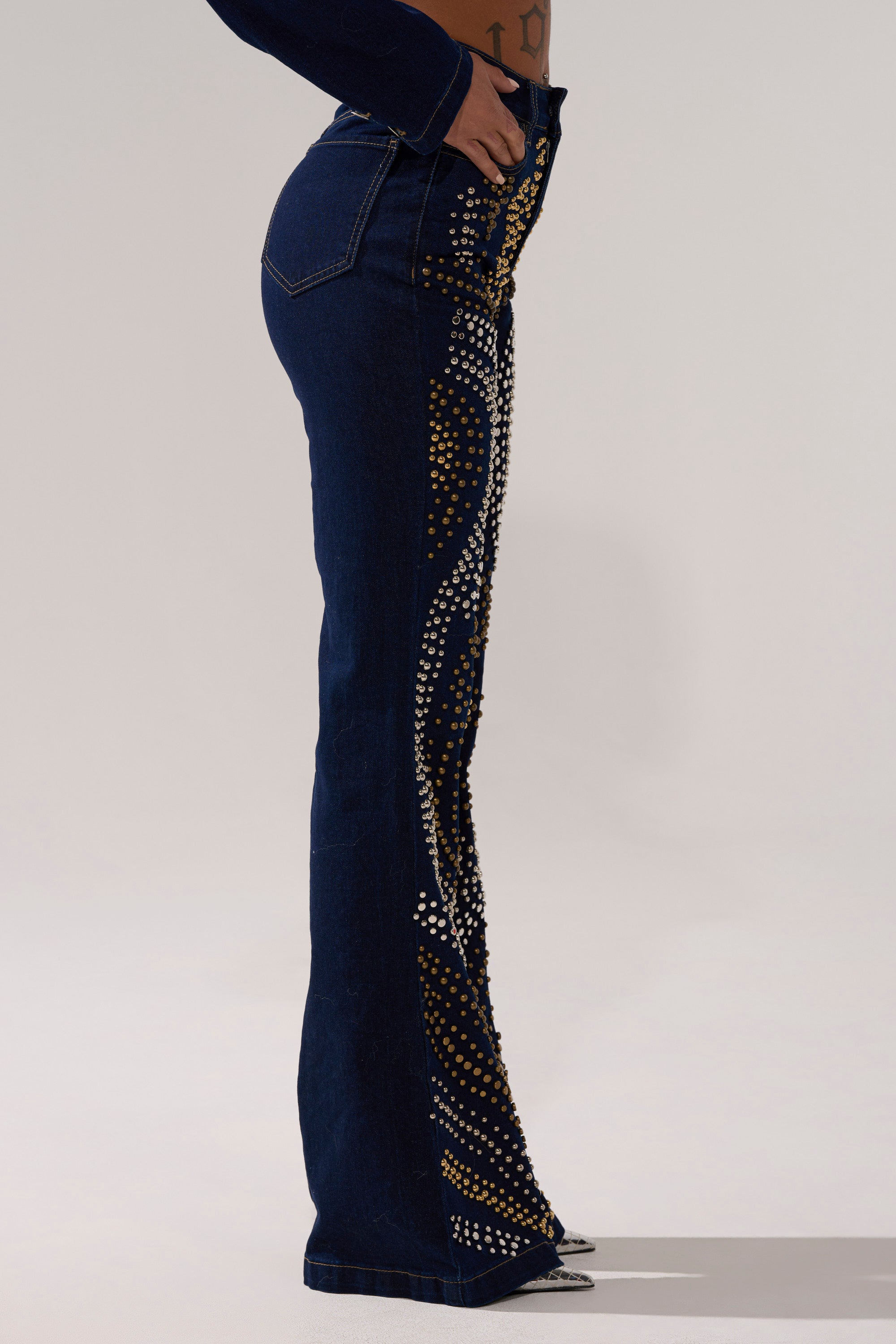 A model wears the CHEYENNE STUDDED FLARE DENIM, dark blue high-rise jeans with a fitted silhouette and gold, silver, and white rhinestone patterns along the side and front, standing out against a plain light gray background.