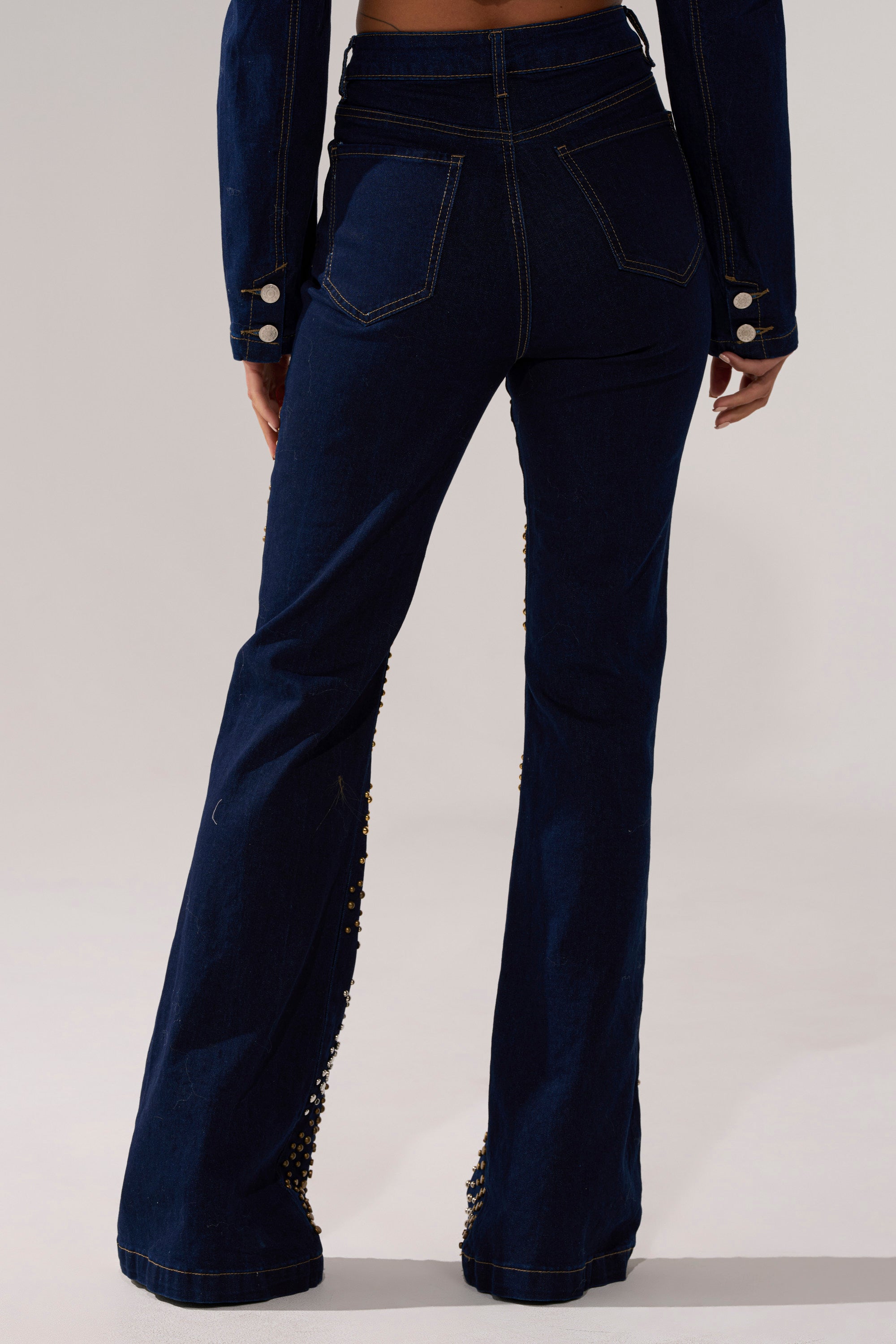 A person faces away, wearing CHEYENNE STUDDED FLARE DENIM—dark blue, high-rise jeans with contrast stitching and gold studs—paired with a matching jacket featuring decorative sleeve buttons.