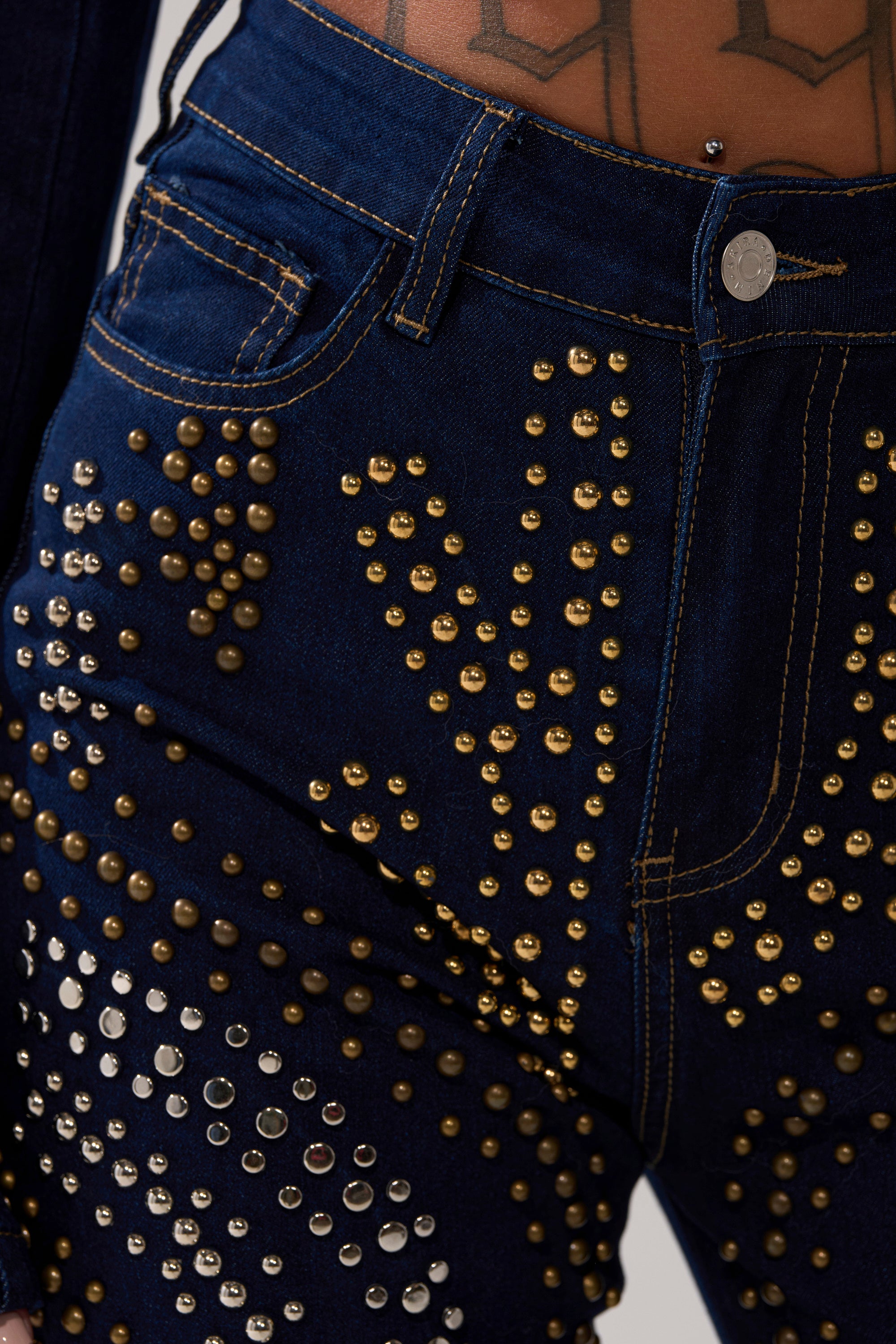 Close-up of a person wearing the CHEYENNE STUDDED FLARE DENIM, dark blue high-rise jeans adorned with gold and silver stud patterns. Waistband sits below a visible pierced, tattooed lower abdomen.