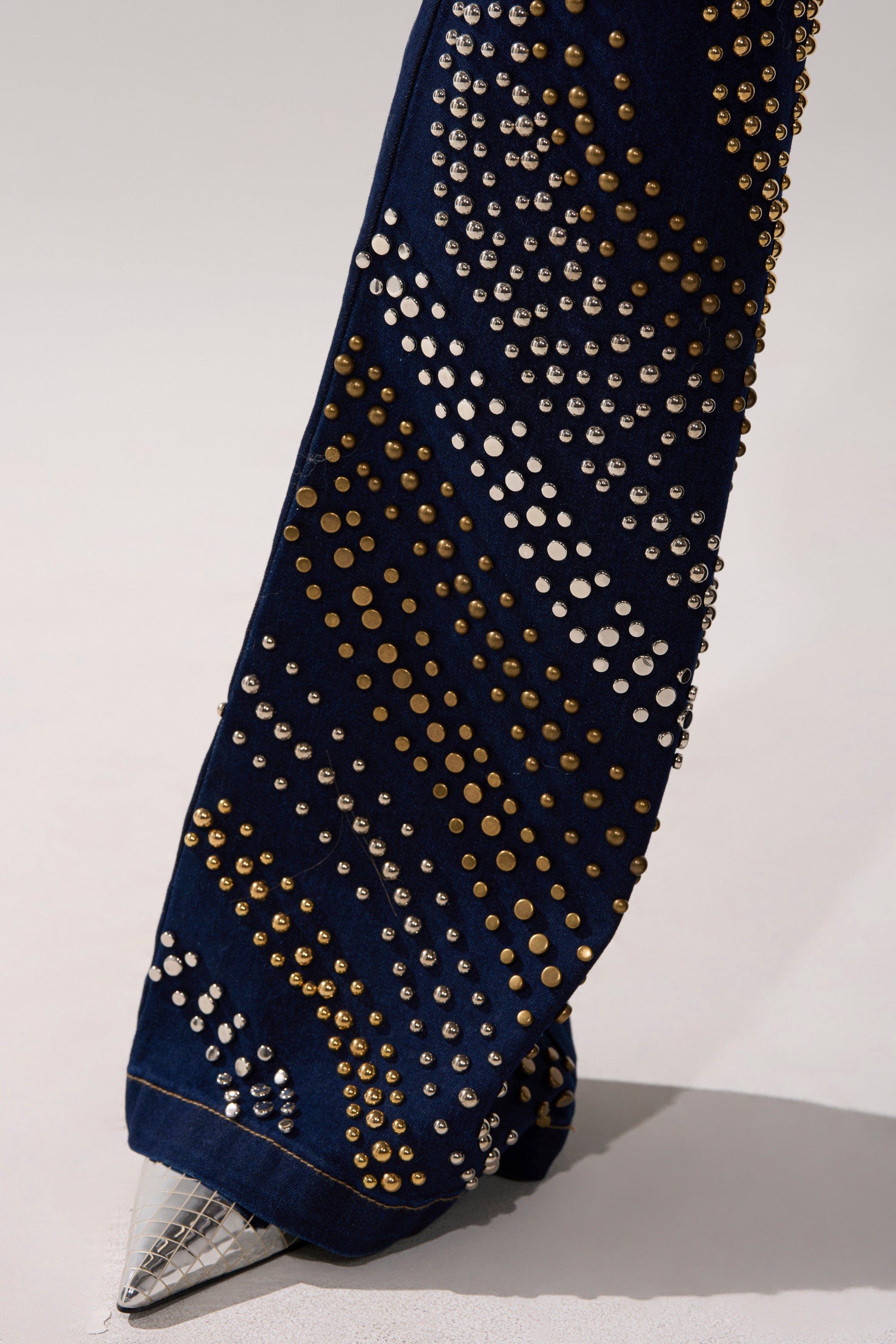 A close-up of the CHEYENNE STUDDED FLARE DENIM reveals dark fabric with gold and silver studs in geometric rows. The hem drapes stylishly over a pointed-toe silver shoe.