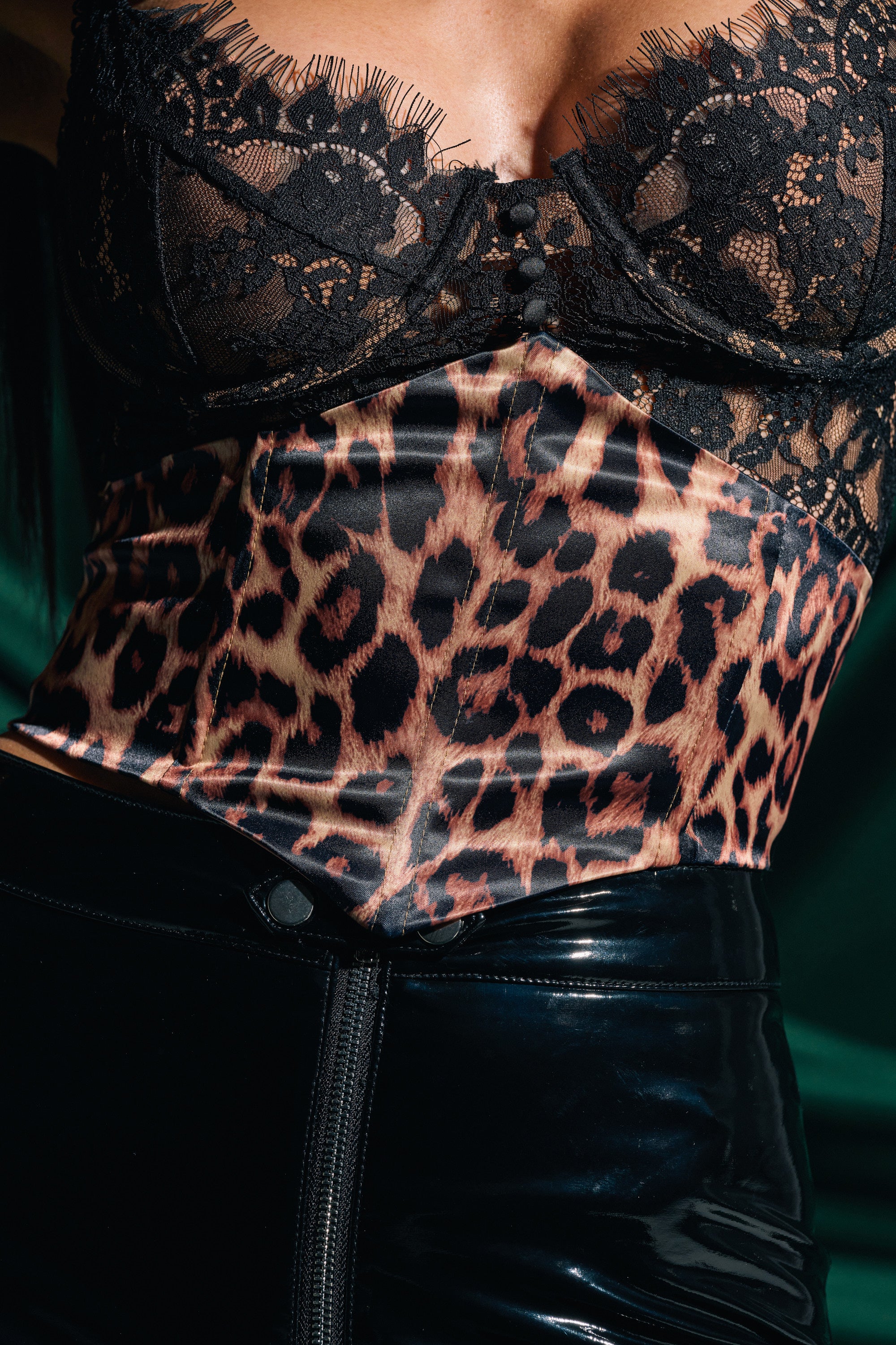 A close-up of a person wearing a black lace top, the IN THE WILD SATIN CHEETAH BELT over a leopard print corset, and shiny black pants against a dark green background.
