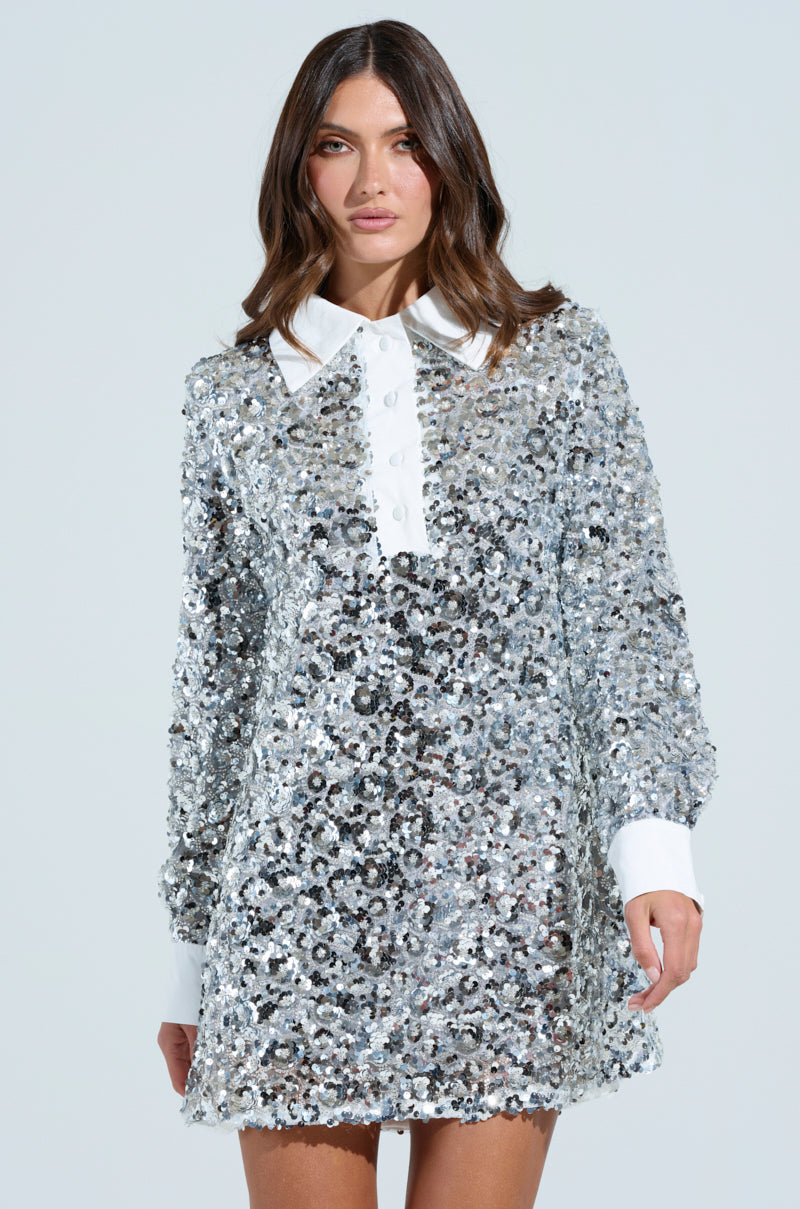 A woman wearing the SO POSH BEADED SEQUIN SHIRT DRESS, featuring a mesh base, sparkly sequins, a white collar and cuffs, stands against a plain background. Her long-sleeved dress complements her wavy brown hair as she gazes directly into the camera.
