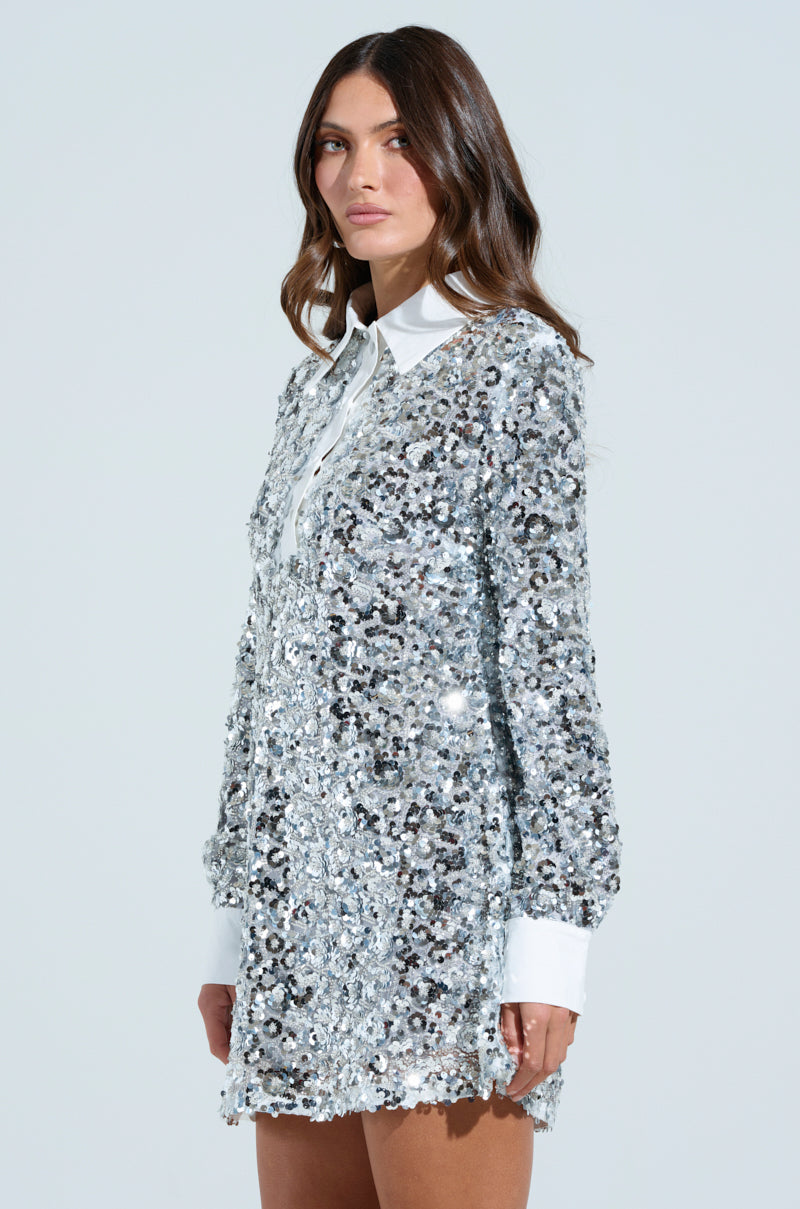 A woman with long brown hair stands against a plain background in a dazzling SO POSH BEADED SEQUIN SHIRT DRESS, featuring a glistening silver design with a white collar and cuffs, its mesh base adorned with intricate sequin and bead embellishments.