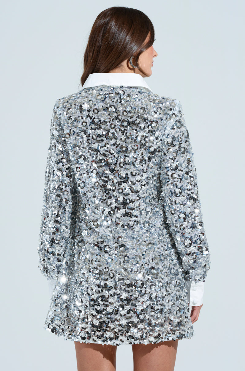 A person with long brown hair faces away, wearing the SO POSH Beaded Sequin Shirt Dress, which features sparkling silver embellishments and a white collar, set against a plain light blue background.