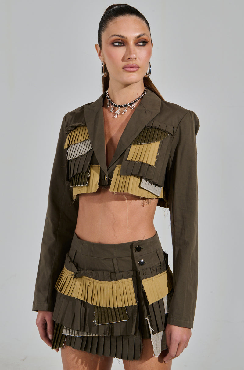 A woman confidently poses in a unique ensemble featuring the "Let Me Down Slowly Cropped Blazer in Olive" and a patchwork skirt in olive green and beige. She flaunts a sleek ponytail, bold makeup, and a statement necklace.