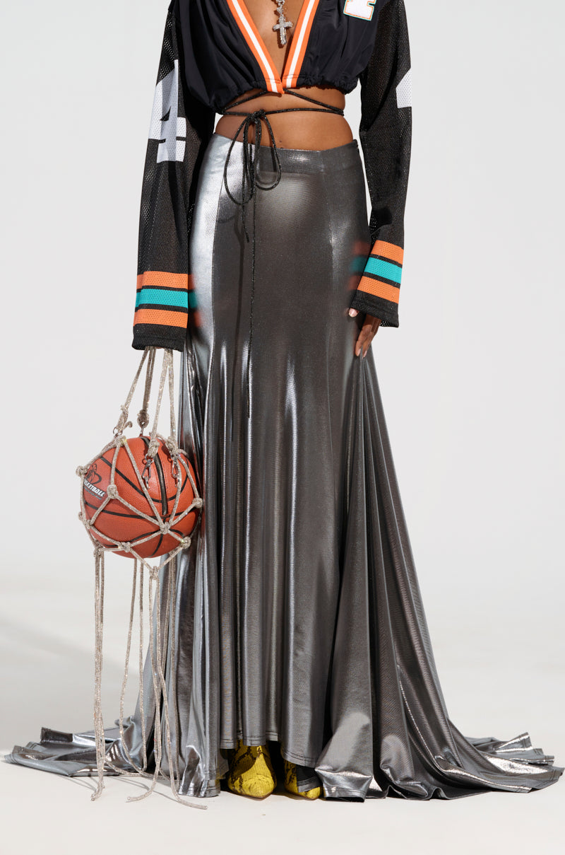 A person in a sporty outfit features a black crop top with multi-colored sleeves and a curve-skimming fit. The SHAKE IT UP MAXI SKIRT in metallic silver adds an edgy twist as they hold a basketball wrapped with ropes. High-low design yellow shoes add contrast to the ensemble.