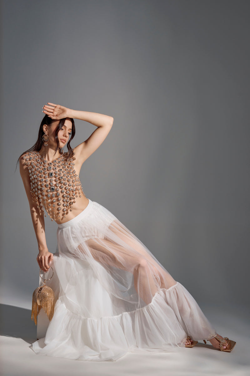 Under soft lighting against a neutral gray background, a woman poses in the TRY ME MESH MAXI SKIRT IN WHITE with a beaded top and gold fringe handbag, raising one arm to her forehead and casting a shadow over her face.