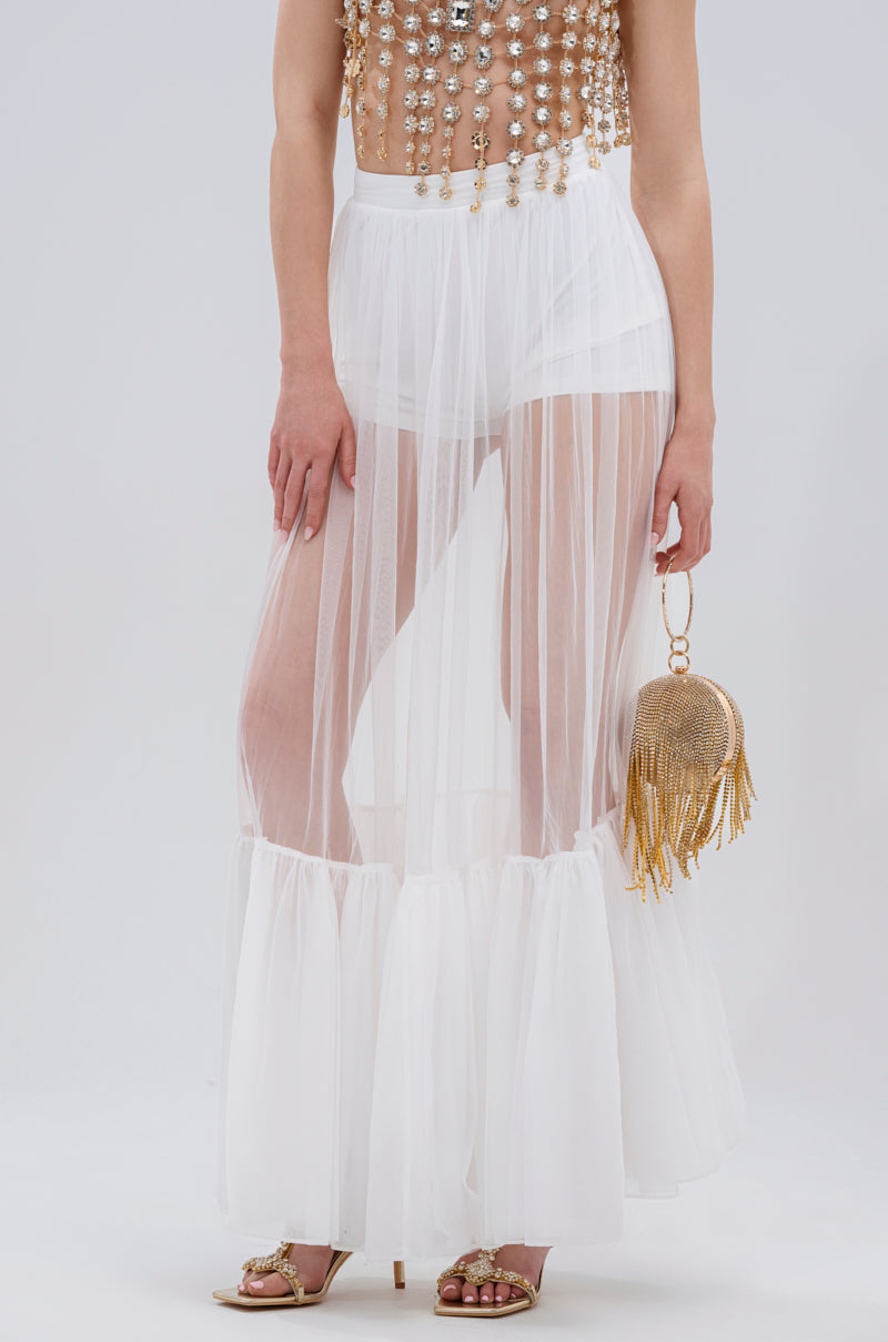 Wearing the TRY ME MESH MAXI SKIRT IN WHITE as a high-waist sheer layer over white shorts, gold sandals, and a gold beaded top, she pairs it with a fringe sparkly gold handbag. The chic look pops against the plain light gray background.