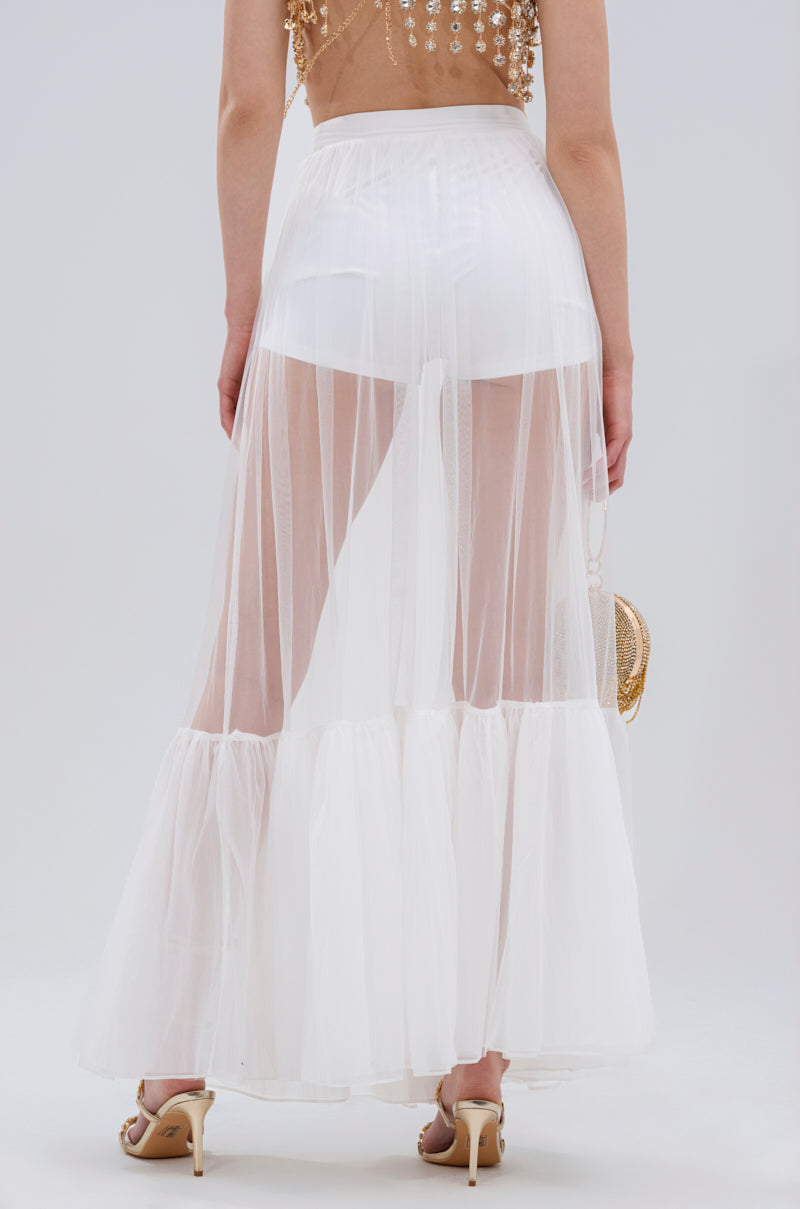A person is seen from behind wearing the TRY ME MESH MAXI SKIRT IN WHITE with ruffle detailing over white shorts, nude high-heeled sandals, and carrying a small round handbag.
