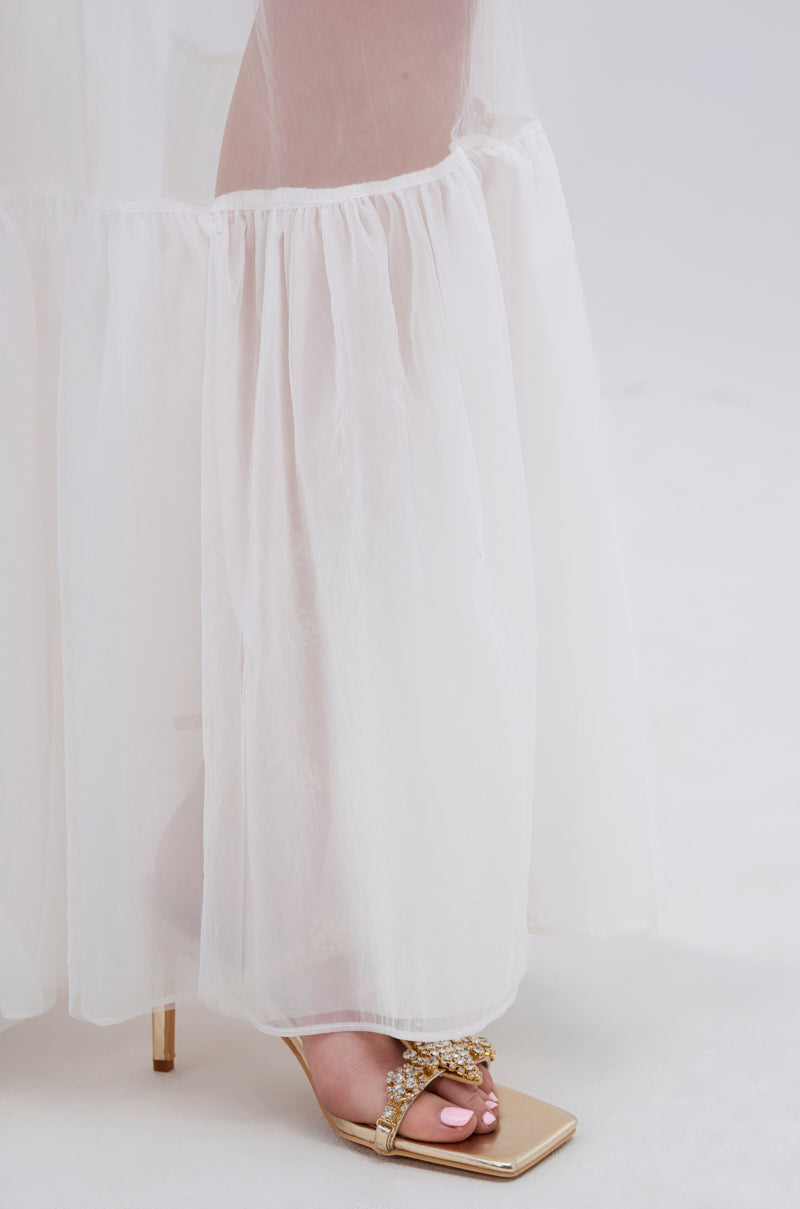 A close-up of the TRY ME MESH MAXI SKIRT IN WHITE brushing over a woman's foot, paired with an elegant gold heeled sandal featuring decorative details against a soft, light background.