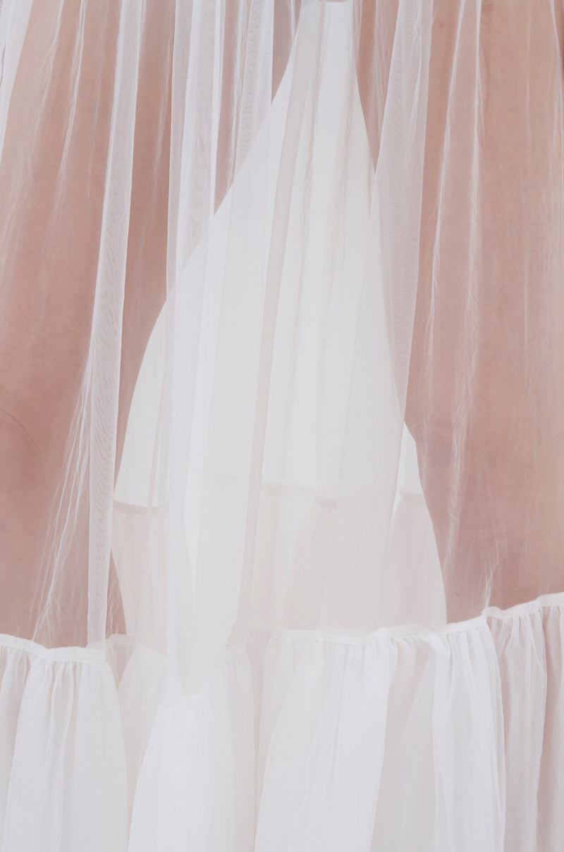 A close-up of the TRY ME MESH MAXI SKIRT IN WHITE, with sheer mesh fabric draped and gathered, soft natural light revealing faint outlines of legs—a subtle glimpse through the stylish maxi skirt.