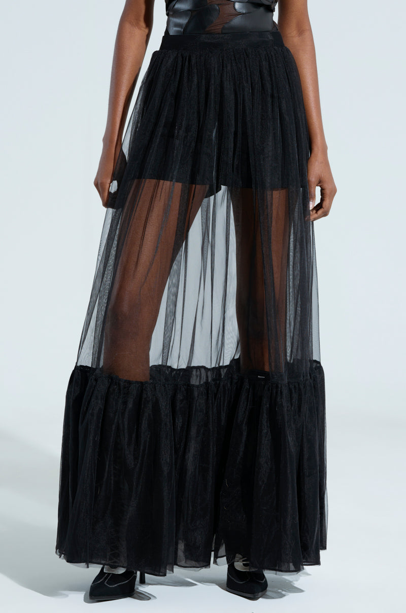 A model poses in the TRY ME MAXI SKIRT, featuring a sheer black tulle design with ruffled construction and gathered hem detailing. The maxi skirt is styled with black shorts underneath and complemented by black shoes, against a light and minimalistic background.