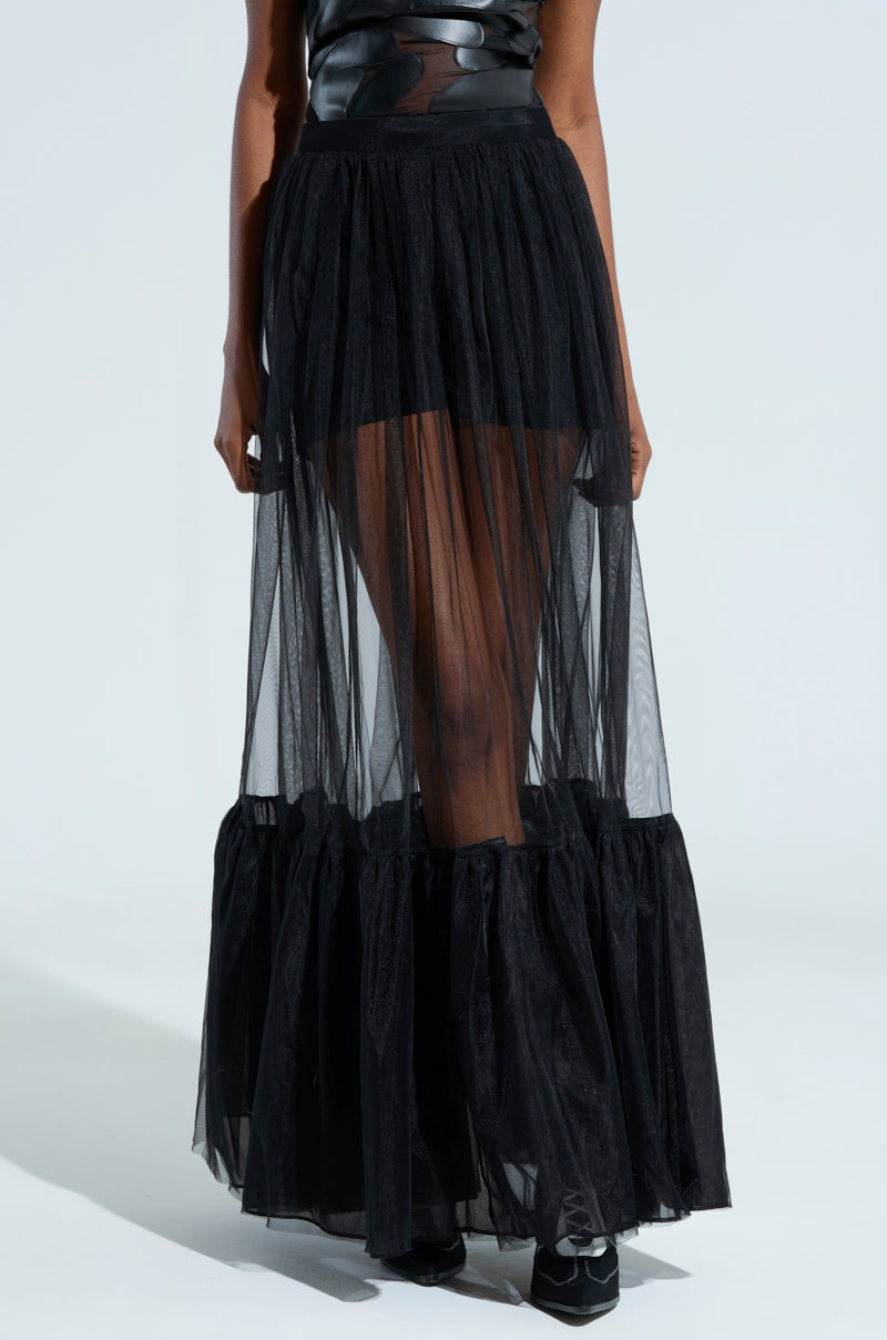 A person wearing the TRY ME MAXI SKIRT, a black sheer maxi skirt featuring tulle fabrication and a ruffled hem over a shorter opaque skirt. Complemented by black footwear, the ensemble combines various textures for a fashionable, layered look.