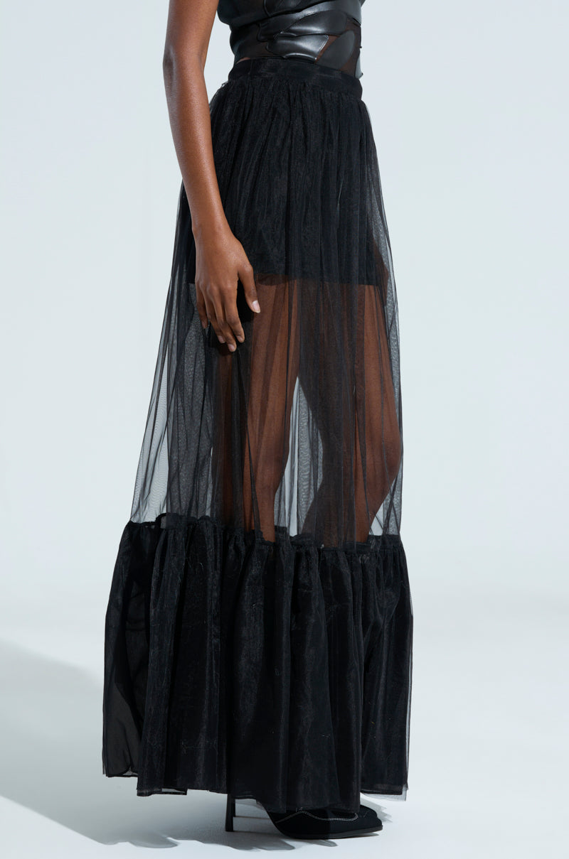 A person wearing the stylish TRY ME MAXI SKIRT, which features a black sheer design with elegant ruffled construction over a shorter black skirt. The outfit is complemented by a fitted black top, and the person is standing against a light background.