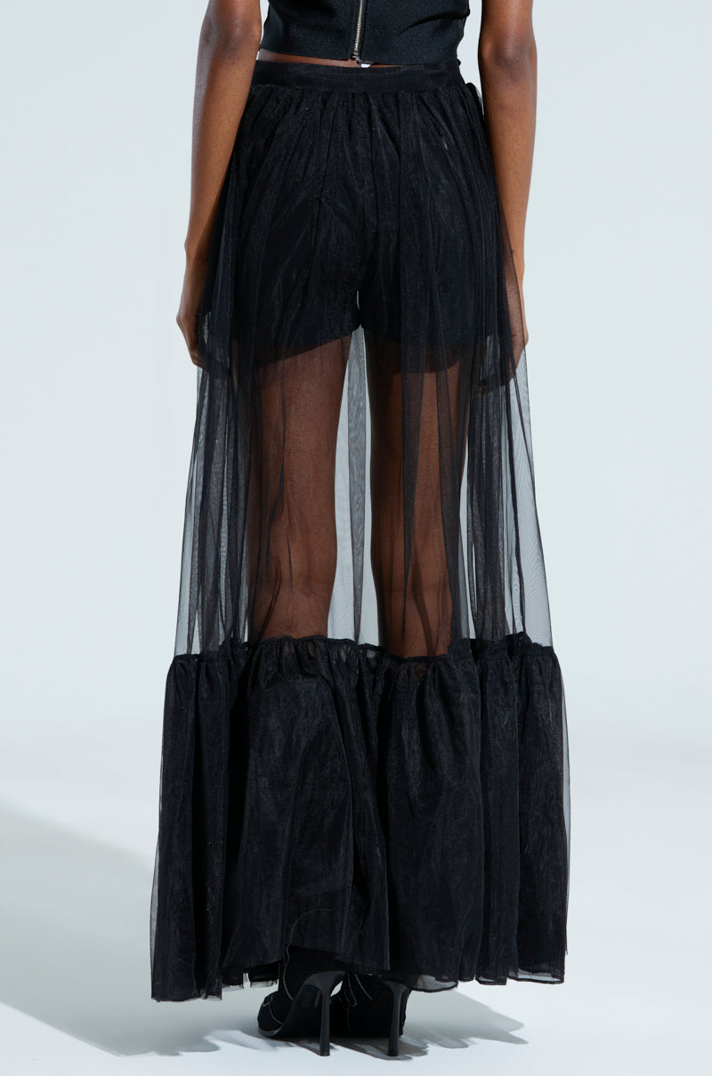 A person is wearing the TRY ME MAXI SKIRT, a black tulle maxi skirt that is sheer from the waist down to the ankles. Black shorts peek through underneath and are paired with black boots. The background is a plain, light color.