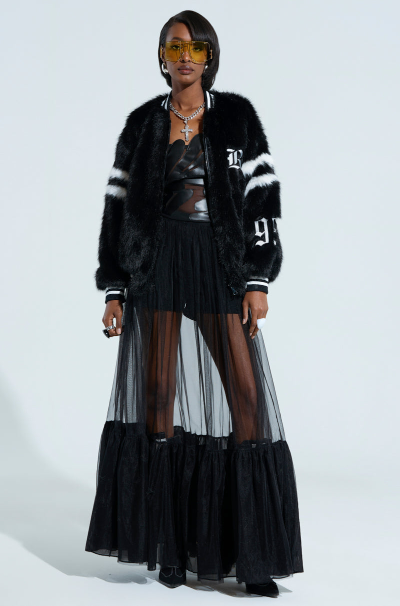 A person wearing a black fur jacket, a strapless leather top, and the TRY ME MAXI SKIRT in sheer black fabric stands confidently. They have short hair and are adorned with yellow-tinted sunglasses and a necklace. The plain white background accentuates the subtle elegance of their tulle fabrication.