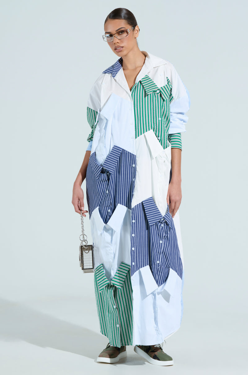 An individual wearing the NOT YOUR BOYFRIENDS SHIRT MAXI DRESS, composed of mixed striped fabric in blue, white, and green like dress shirts. They hold a small metallic handbag and wear glasses, with hair styled back against a light blue background.