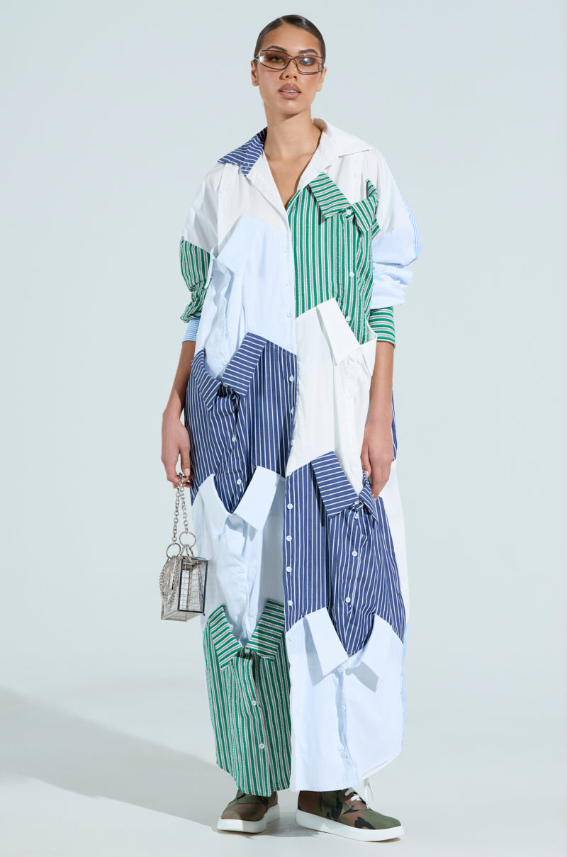 A model displays the "NOT YOUR BOYFRIEND'S SHIRT" maxi dress featuring green, blue, and white vertical stripes. She accessorizes with a small transparent handbag and sunglasses, while her casual shoes blend with the minimalist backdrop.