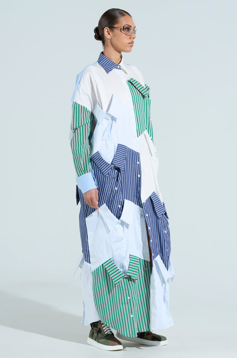 A person in glasses stands against a light background, wearing NOT YOUR BOYFRIEND'S SHIRT MAXI DRESS with an oversized silhouette. The outfit combines eclectic stripes and plain sections in blue, green, and white tones, complemented by matching sneakers.
