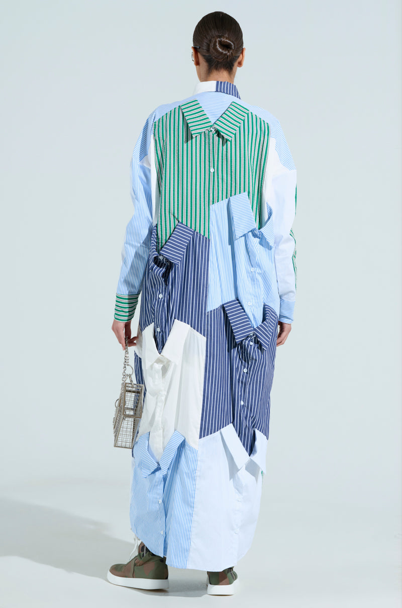 Facing away from the camera, an individual wears a poplin patchwork NOT YOUR BOYFRIENDS SHIRT MAXI DRESS with blue, white, and green stripes. Complemented by a small handbag and green sneakers, their tied-back hair accentuates the ensemble's oversized silhouette.