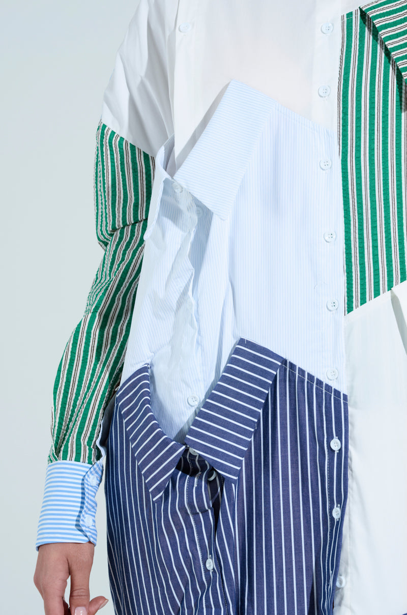 A person is clad in a long-sleeve, oversized NOT YOUR BOYFRIENDS SHIRT MAXI DRESS featuring vertical patchwork stripes in green, blue, white, and navy on a light blue backdrop. It buttons up from neck to thigh.