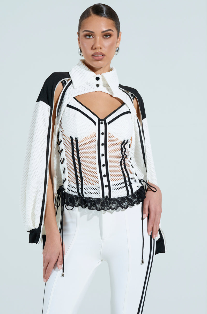 A woman poses confidently in a VIVI JERSEY CORSET, showcasing a stylish black and white ensemble. Her sporty-chic top features perforated jersey mesh with cut-out shoulders and lace-up details, paired with side-stripe pants against a light blue background. 1