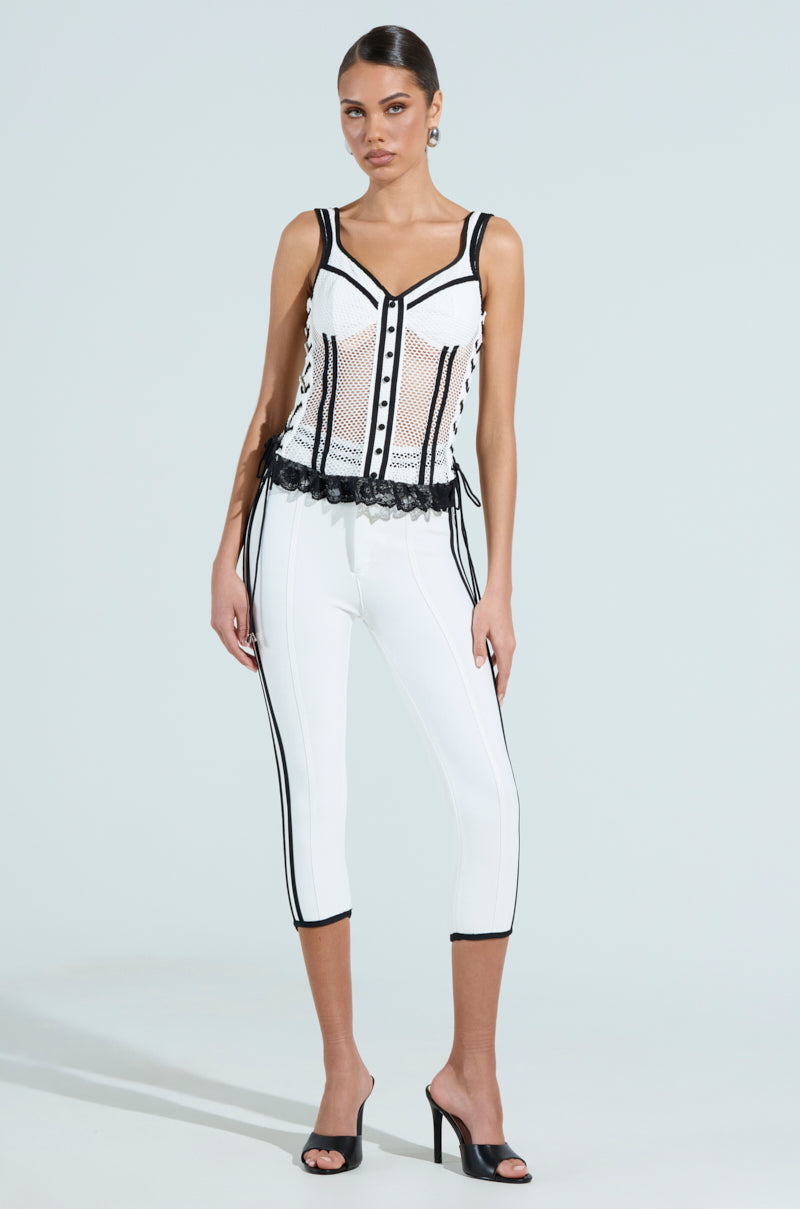 A woman wears a stylish black and white Vivi Jersey Corset with perforated mesh and lace, paired perfectly with white capri pants featuring black stripes and complemented by black high-heeled mules. Her hair is elegantly tied back. 5
