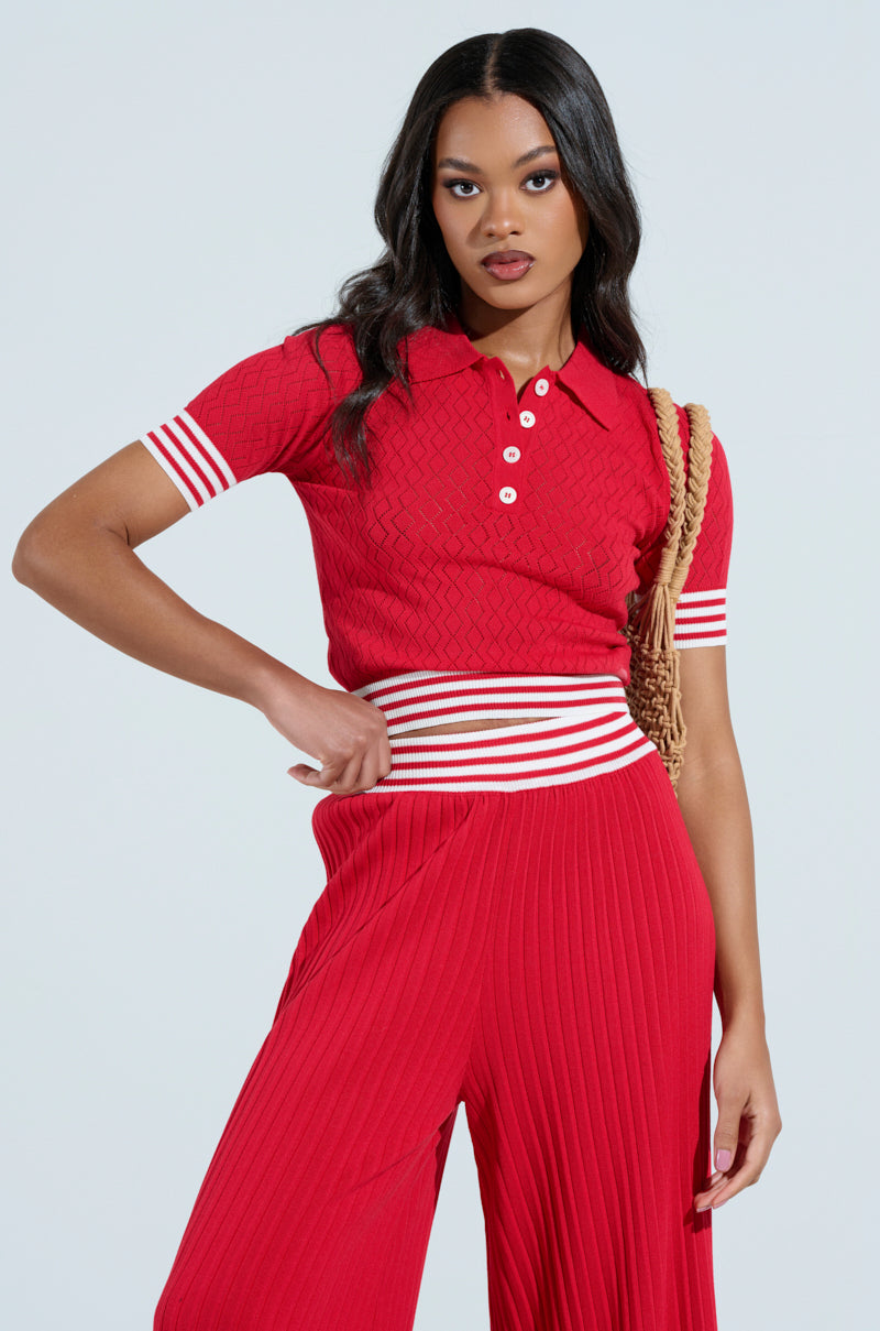 A woman poses confidently in the AKIRA Label GET WITH IT Crop Sweater in red, featuring white buttons and striped sleeves, paired with matching stretchy knit pleated pants. She has a woven bag on her shoulder and long, wavy hair against a simple pale background.