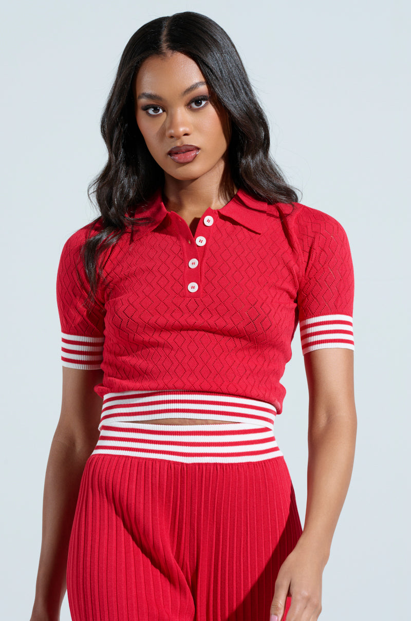 A person in an AKIRA Label GET WITH IT CROP SWEATER, a red short-sleeve knit polo with white buttons and striped cuffs, complements matching stretchy ribbed pants featuring a white-striped waistband against a light gray background.