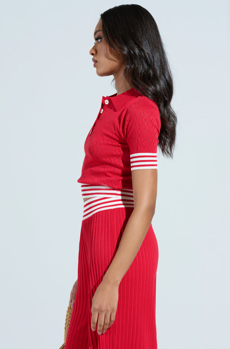 A woman with long black hair stands in profile wearing an AKIRA Label GET WITH IT CROP SWEATER, holding a small straw purse against a plain background, showcasing her chic style with elegance and flair.
