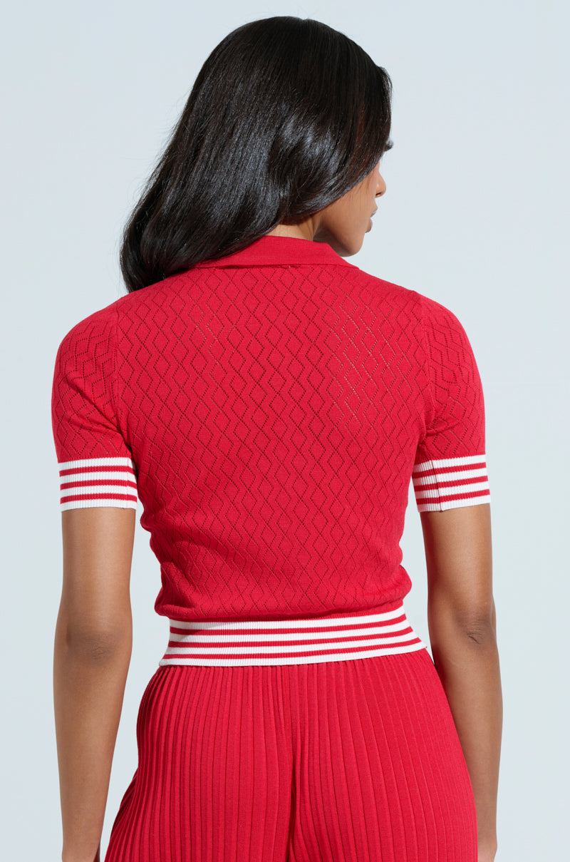 A person with long dark hair, wearing an AKIRA Label GET WITH IT CROP SWEATER in red and matching pants with white stripes at the waist and sleeves, faces away, highlighting the textured knit pattern against a light background.