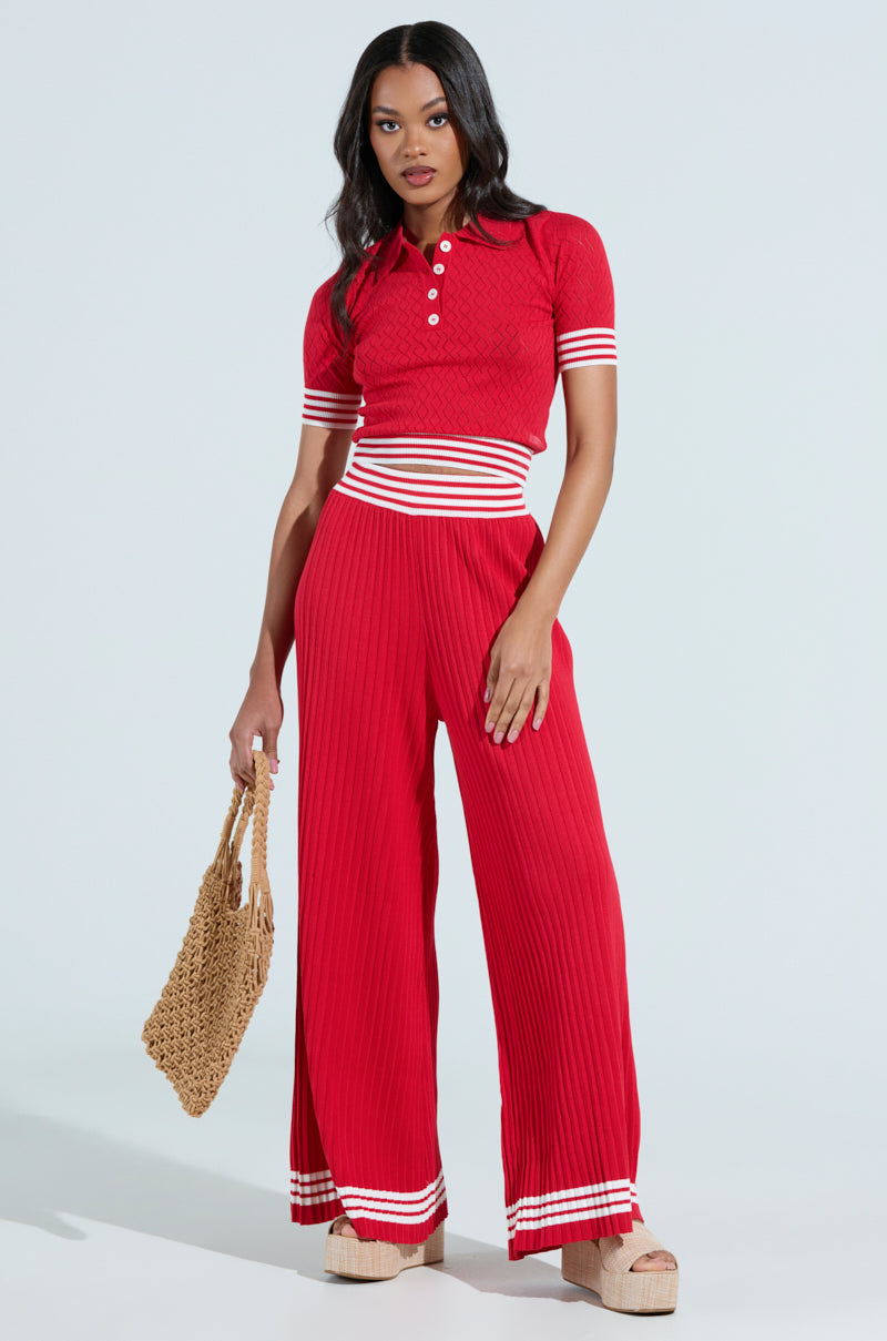A woman confidently poses in a red AKIRA GET WITH IT CROP SWEATER paired with matching wide-leg pants featuring white stripes. She holds a woven bag and wears platform sandals. The stretchy knit outfit delivers a sporty, chic vibe against a neutral backdrop.