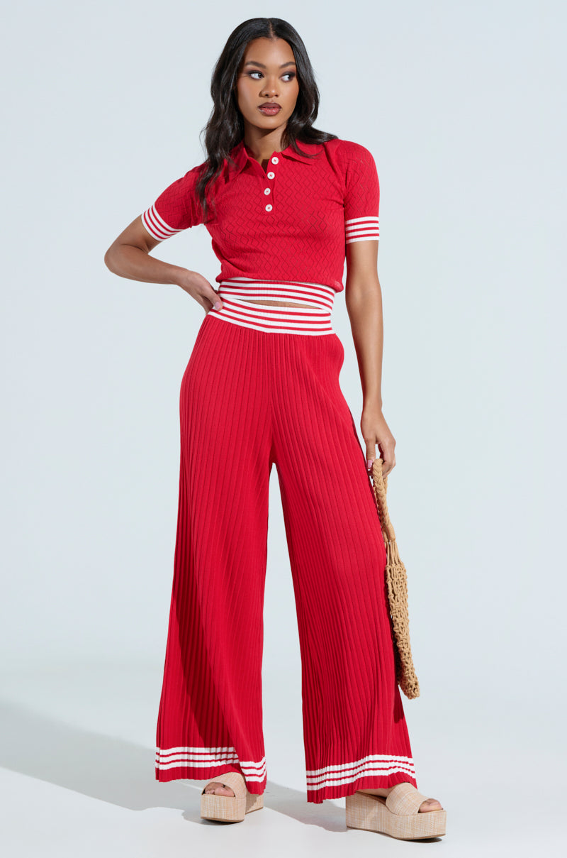 Dressed in a red outfit with white and striped accents, a person stands against a plain background. They're wearing flared pants and the short-sleeve GET WITH IT CROP SWEATER by AKIRA Label, holding a woven bag, and standing in platform sandals.