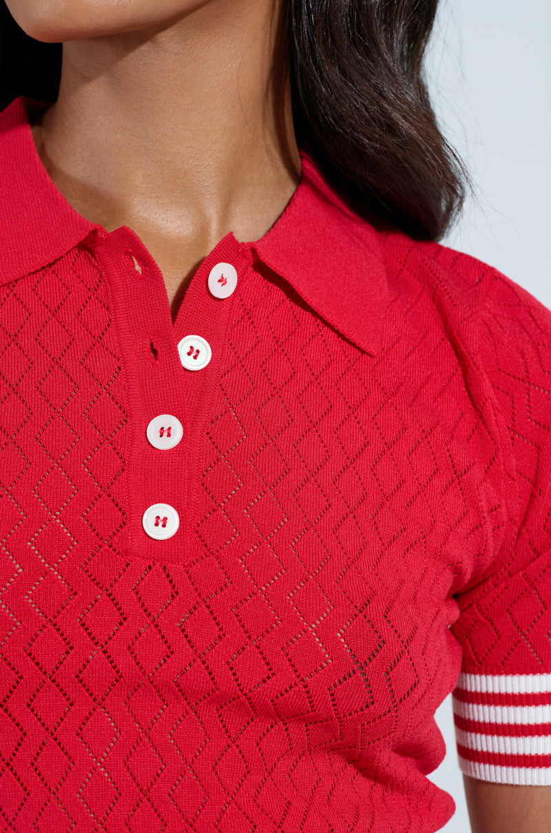 A person wearing the GET WITH IT CROP SWEATER, a red polo shirt with a diamond knit pattern, collar, four white buttons, and white-striped sleeve detail. The background is plain light-colored.