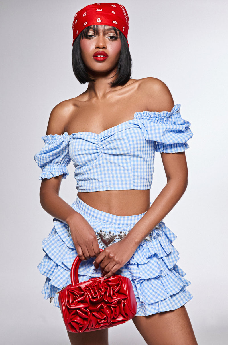 A woman strikes a confident pose in the MORE FUN FOR ME gingham blouse with puff sleeves, paired with a matching ruffled skirt. She's accessorized with a red bandana and handbag, while her bold red lipstick enhances her striking makeup.