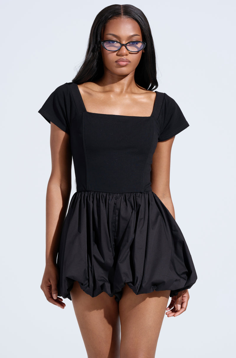 A woman with long straight hair wears blue cat-eye glasses and the KEEP IT SIMPLE BUBBLE ROMPER, featuring a corset-style bodice and bubble hem, standing against a plain light background.