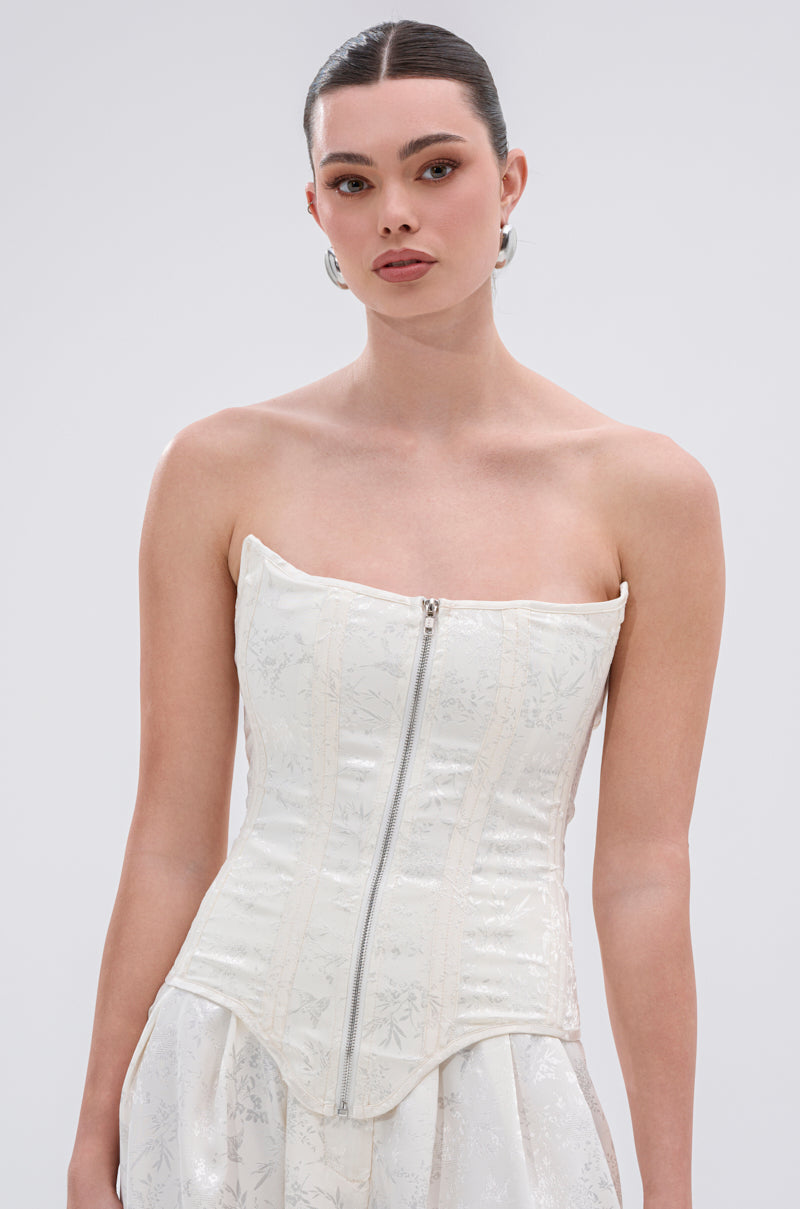 A woman with dark hair in a sleek bun models the DAISY DOVE JACQUARD CORSET, which features structural boning and a front zipper, styled with the matching skirt. She stands against a plain light gray background.