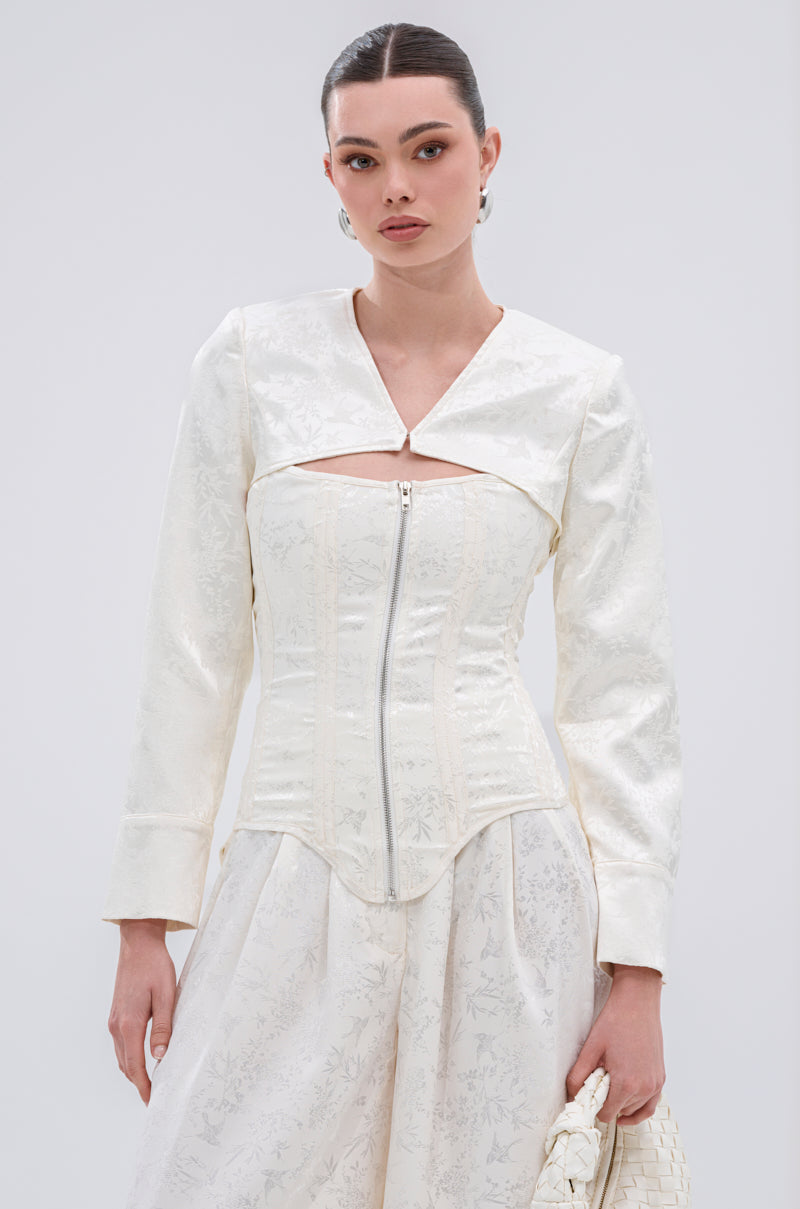 A woman wears the DAISY DOVE JACQUARD CORSET, a white long-sleeve jacket with a cutout neckline and front zipper, over a matching satin skirt. Her hair is tied back with earrings, and she carries a textured white bag.