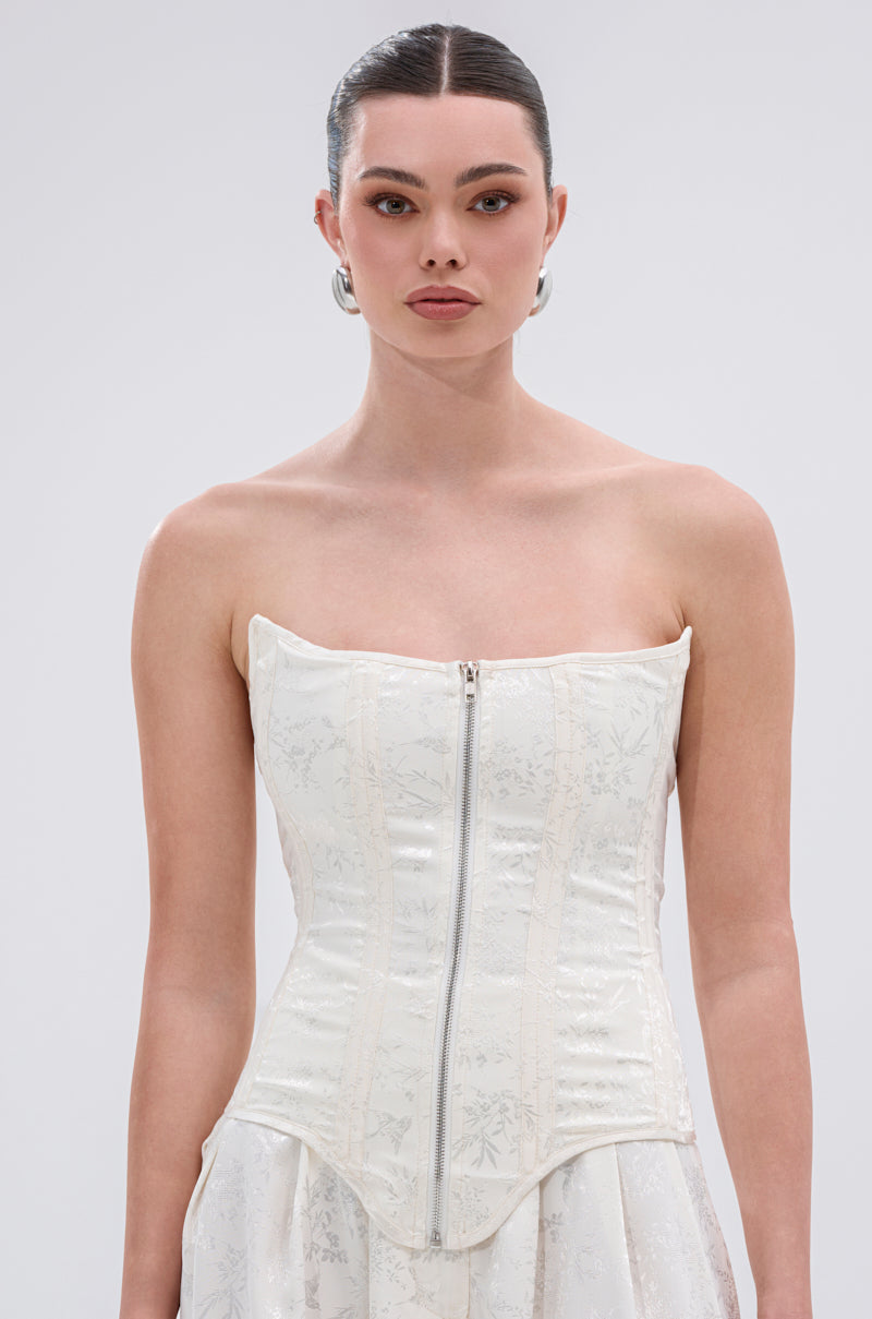 A woman with dark hair tied back wears the DAISY DOVE JACQUARD CORSET, which features structural boning, a front zipper, and subtle floral satin jacquard. She pairs it with a matching skirt and stands against a plain light background.
