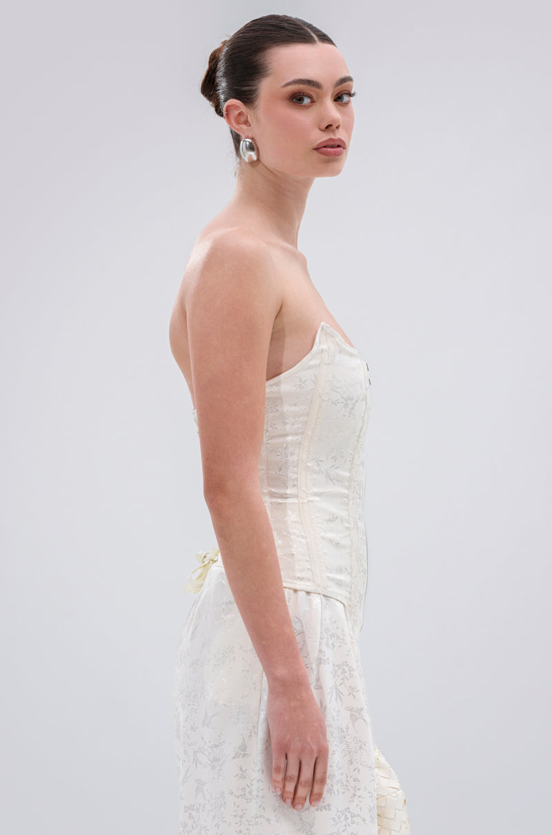 Woman with dark hair in a neat bun stands sideways against a plain light background, wearing the DAISY DOVE JACQUARD CORSET with structural boning and large silver earrings.